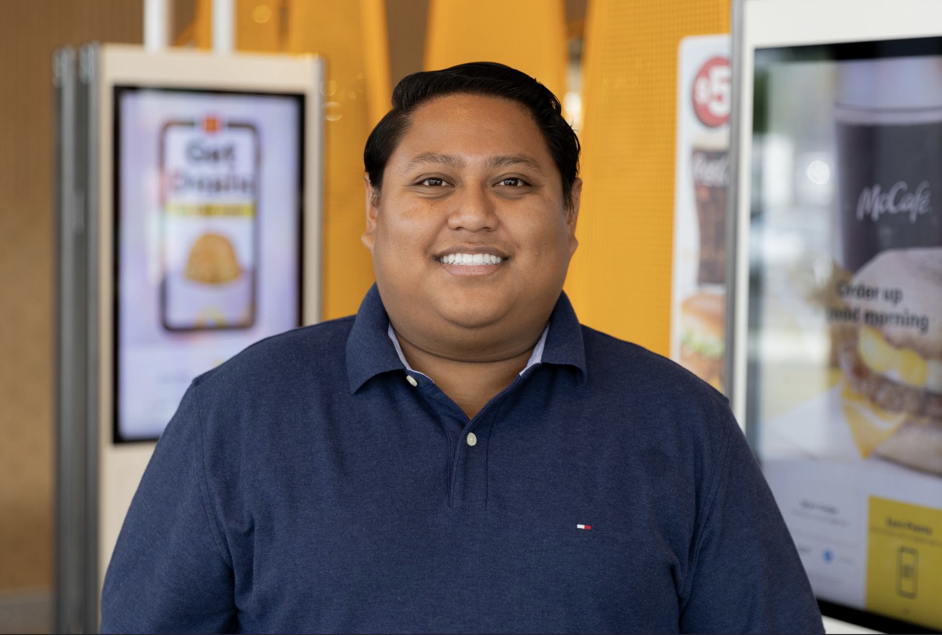 A smiling man with dark hair wearing a navy blue shirt, standing in front of digital menu screens at a fast-food restaurant.