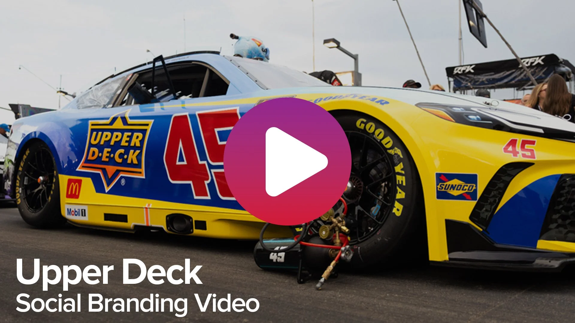 A race car with colorful sponsorship logos and the number 45 is parked on the track, with a large play button overlayed in the center.