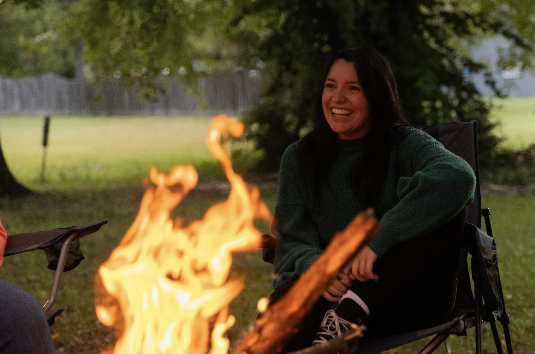 A woman sitting in a camping chair by a campfire outdoors, smiling and enjoying a chilly evening.