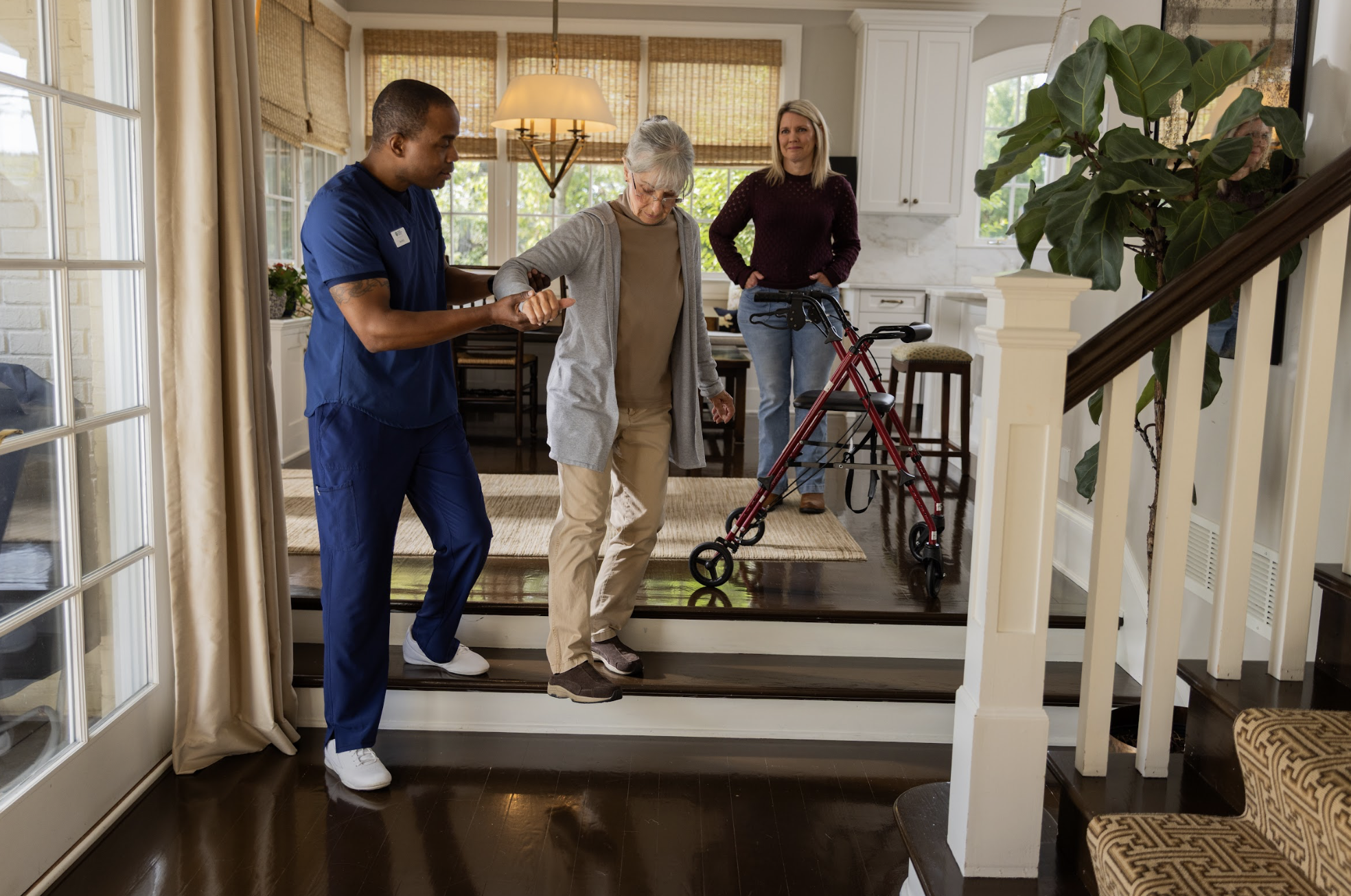 A healthcare assistant supports an elderly woman with a walker as she learns to walk on a small step in her home while a woman observes nearby.
