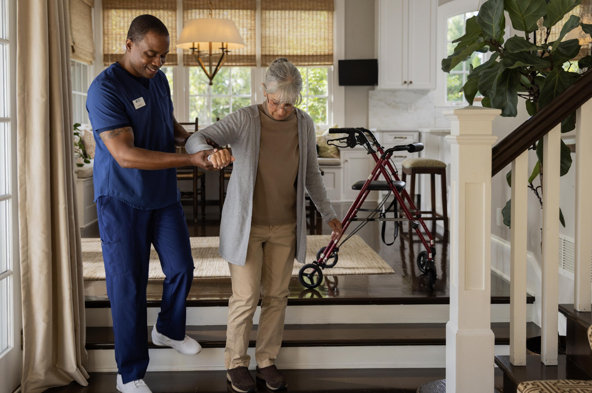 A caregiver assisting an elderly woman with walking using a walker in a home.
