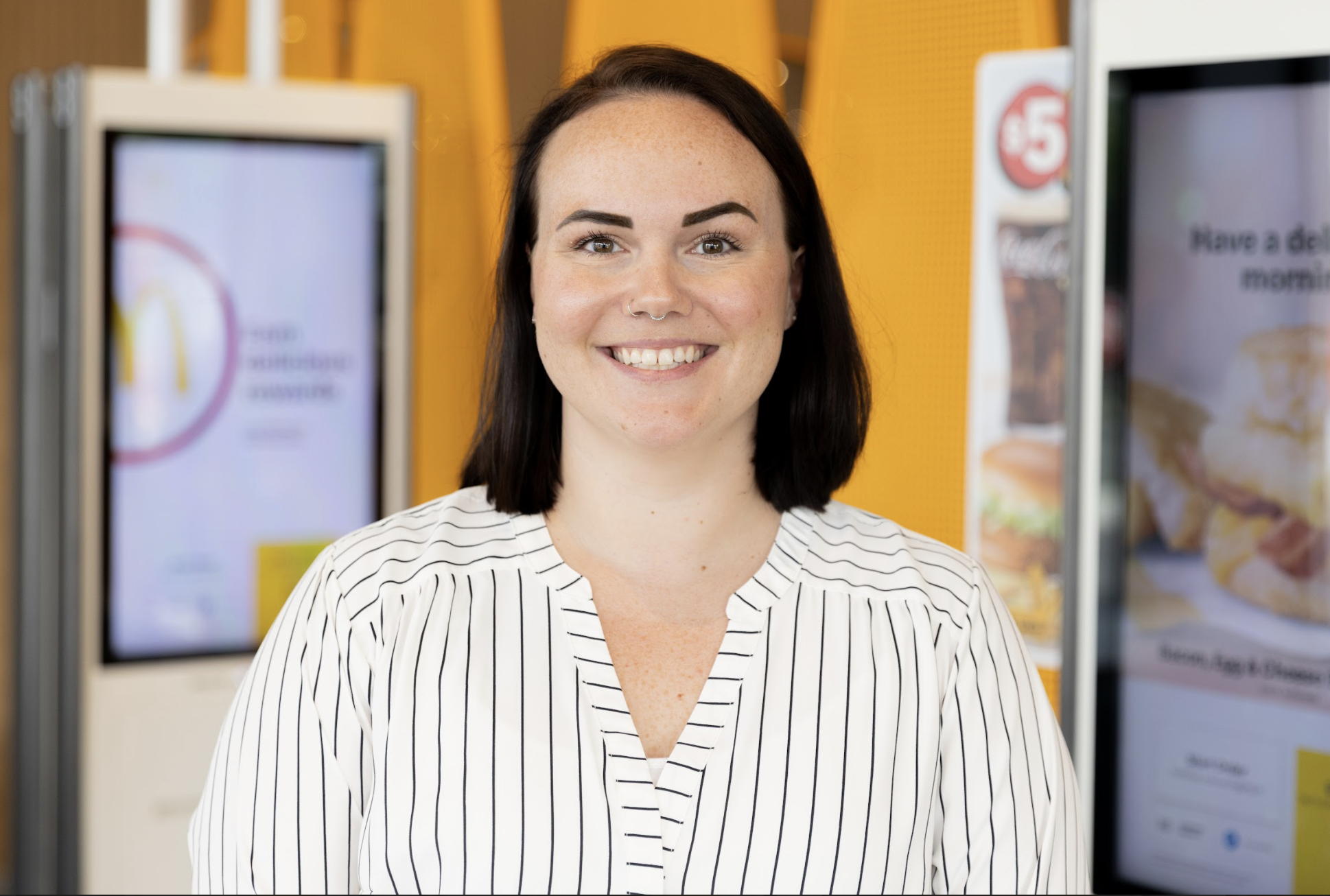 A woman with dark hair, fair skin, and freckles smiling in front of digital screens at a fast food restaurant.