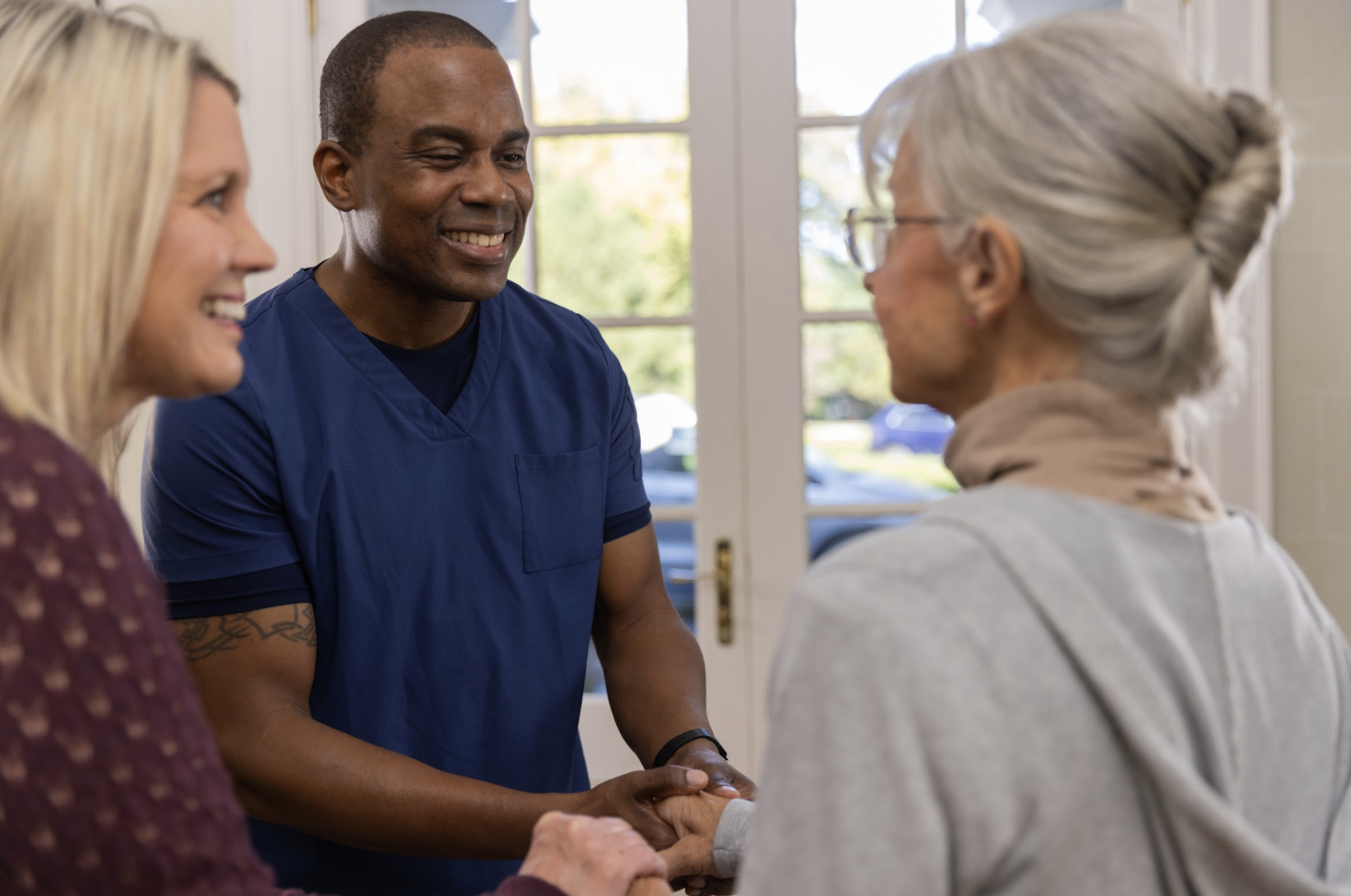 A healthcare worker shaking hands with an elderly woman while an middle-aged woman looks on, inside a well-lit room with glass doors and windows.