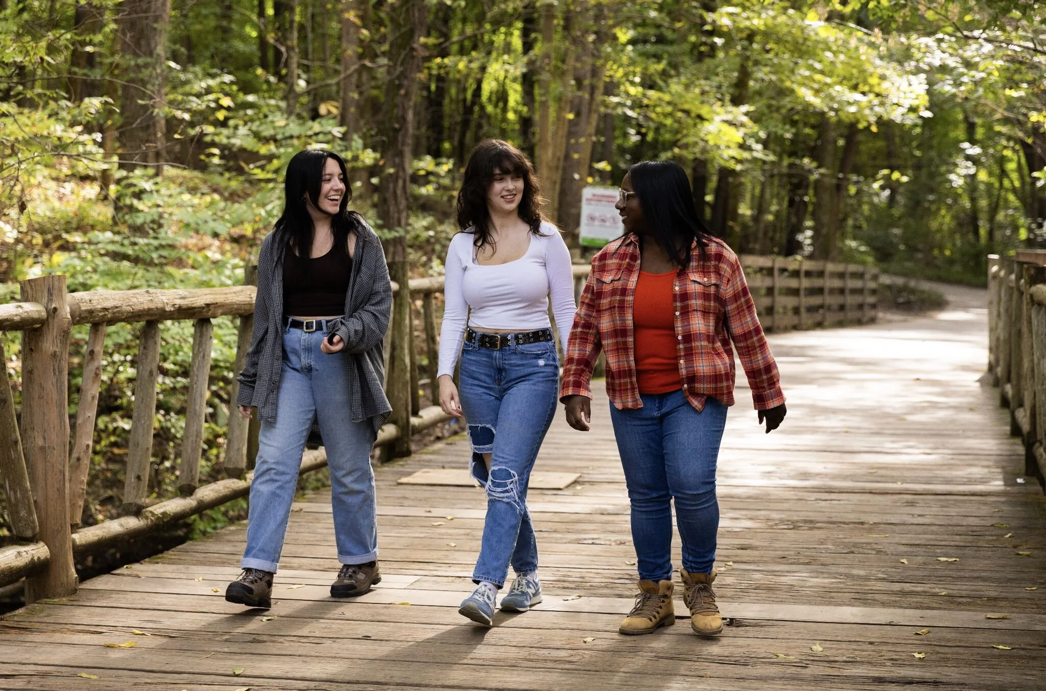 Three women walk and talk on a wooden path through a green forest, smiling and enjoying each other's company.