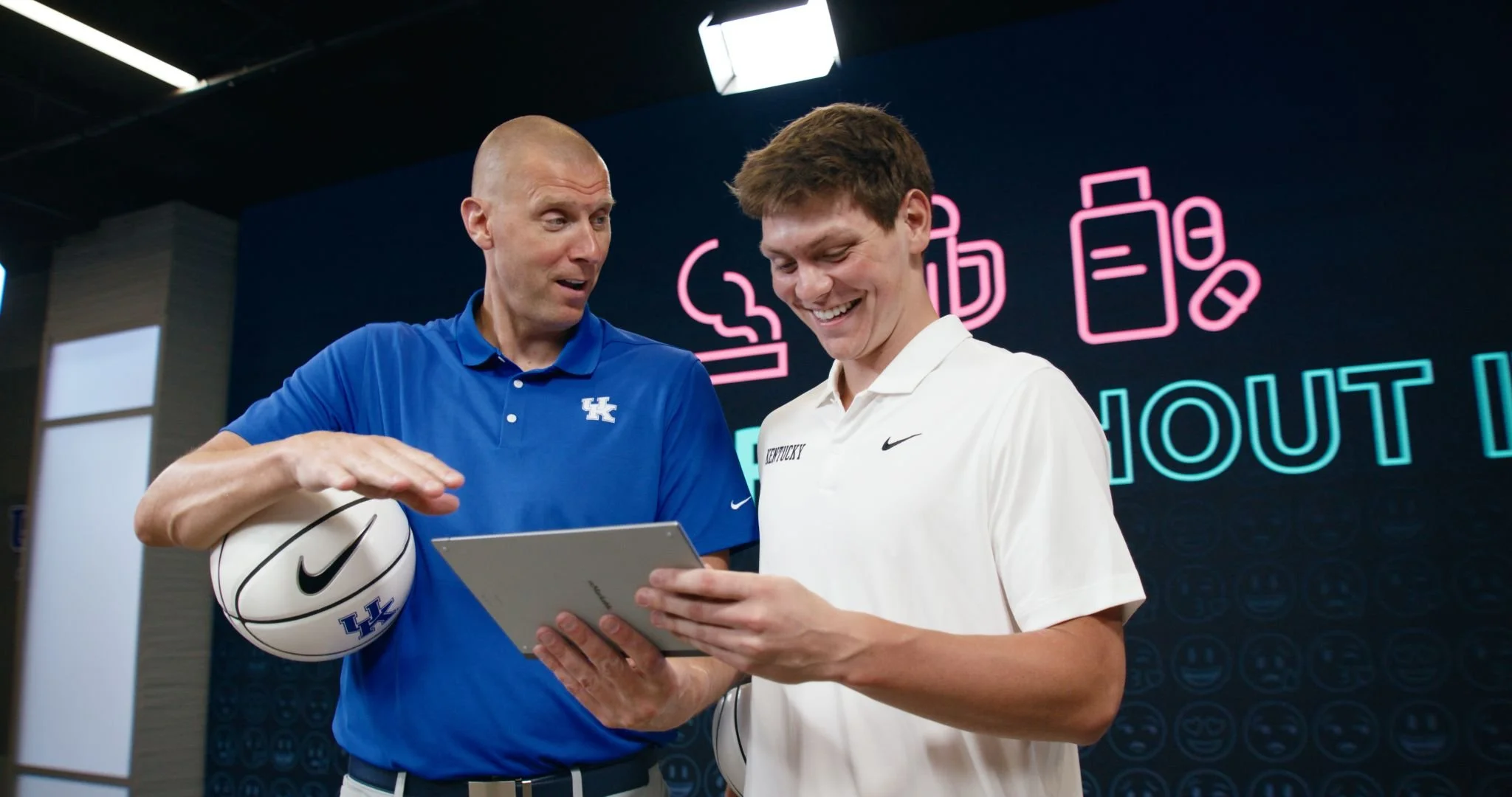 Two men, one in a blue shirt and one in a white shirt, are smiling and looking at a tablet device, with a Kentucky Wildcats logo on his shirt and a basketball with the same logo.