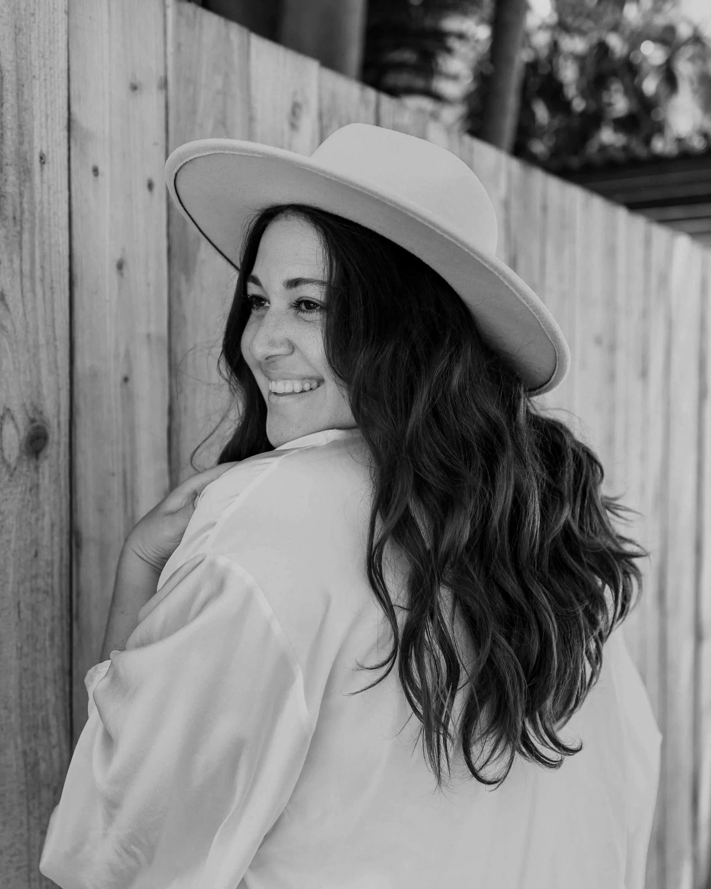 A woman with long wavy hair wearing a wide-brimmed hat and a light-colored shirt, smiling and leaning against a wooden fence outdoors.