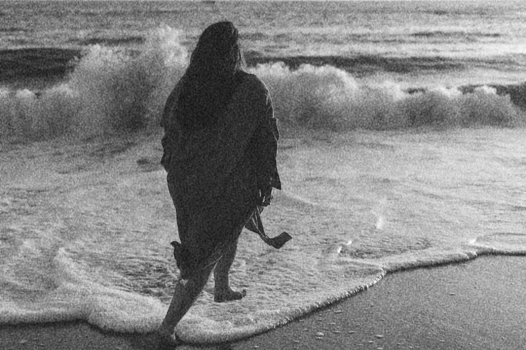 A person with long hair and a jacket walking barefoot on the beach near the water's edge, holding a pair of shoes, during sunset or sunrise.