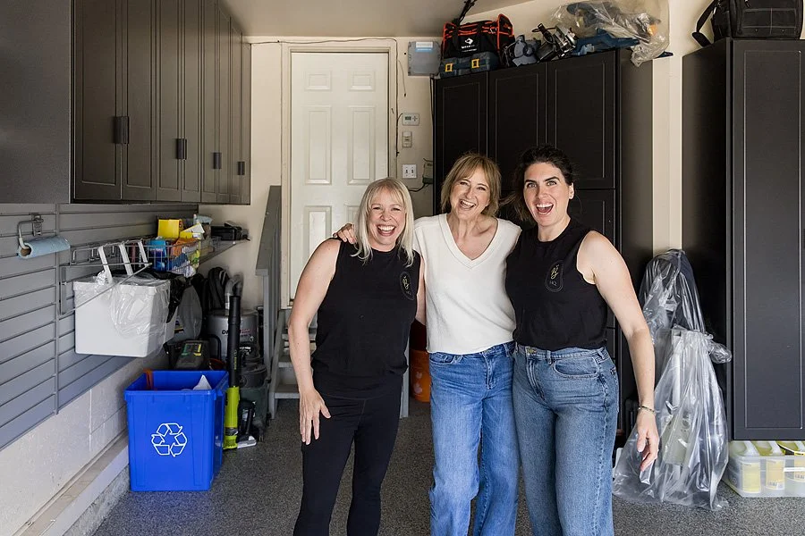 Three women standing together inside a garage, smiling and laughing. Two women are wearing black sleeveless tops and jeans, and the woman in the middle is wearing a white sweater and blue jeans.