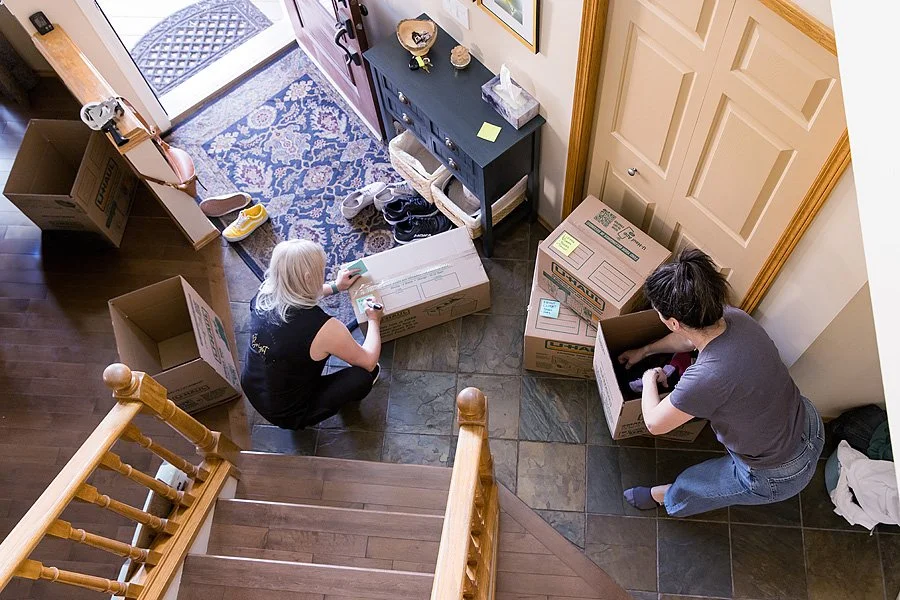 Two women packing boxes near the entrance of a house, with shoes and a patterned rug nearby.