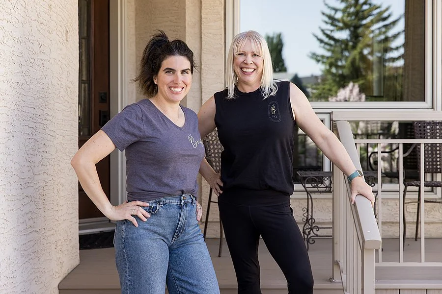 Two women standing on a porch smiling at the camera, with one resting her hand on her hip and the other holding onto the porch railing. The woman on the left has dark hair tied back, wears a gray T-shirt and blue jeans. The woman on the right has blonde hair, wears a black sleeveless top and black pants.