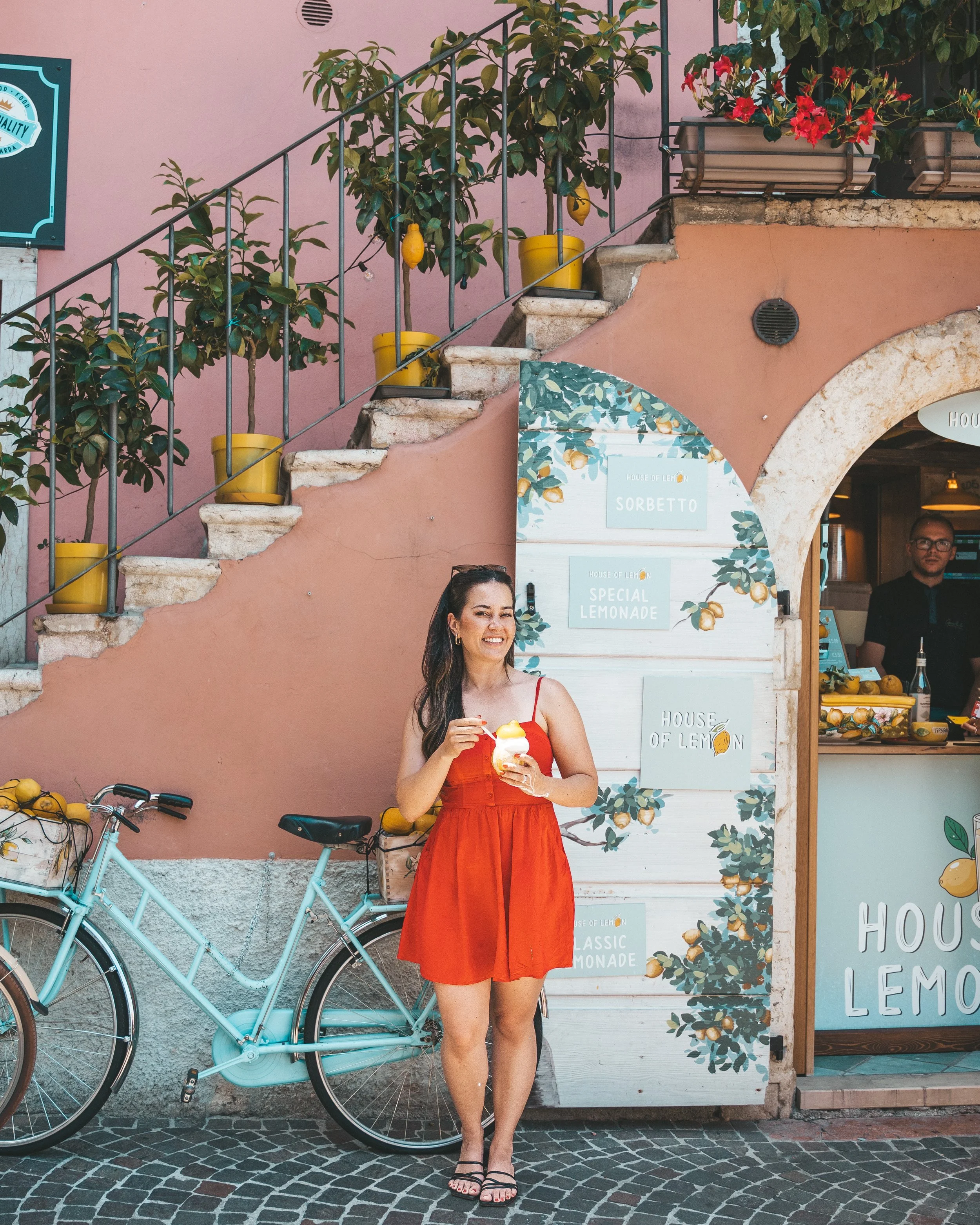 Woman in sundress enjoying a fresh lemon sorbet outside House of Lemonade shop with a vintage blue bicycle in Limone sul Garda