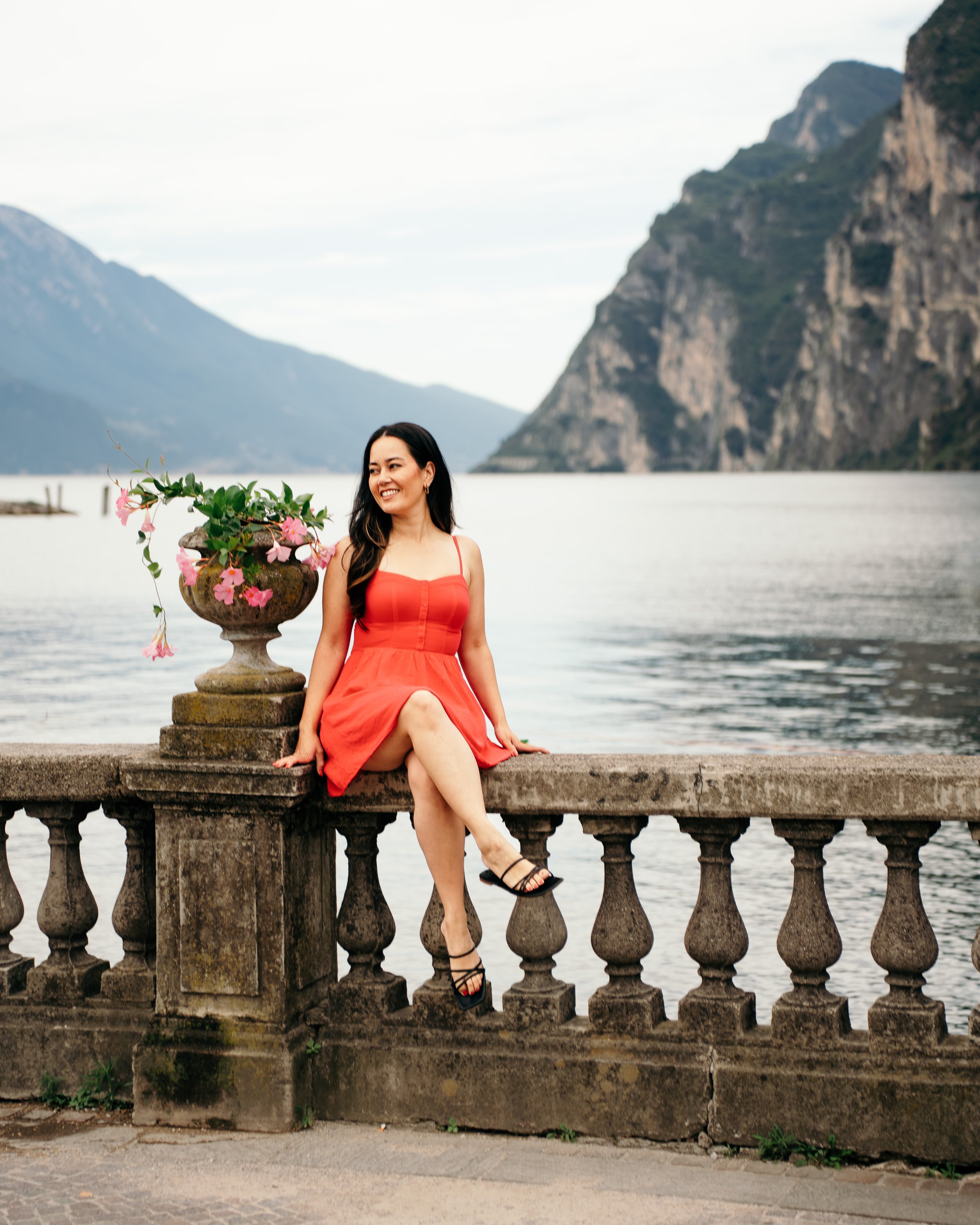 A woman in a sundress sitting on a stone balcony with floral urns overlooking Riva del Garda and mountains in Italy