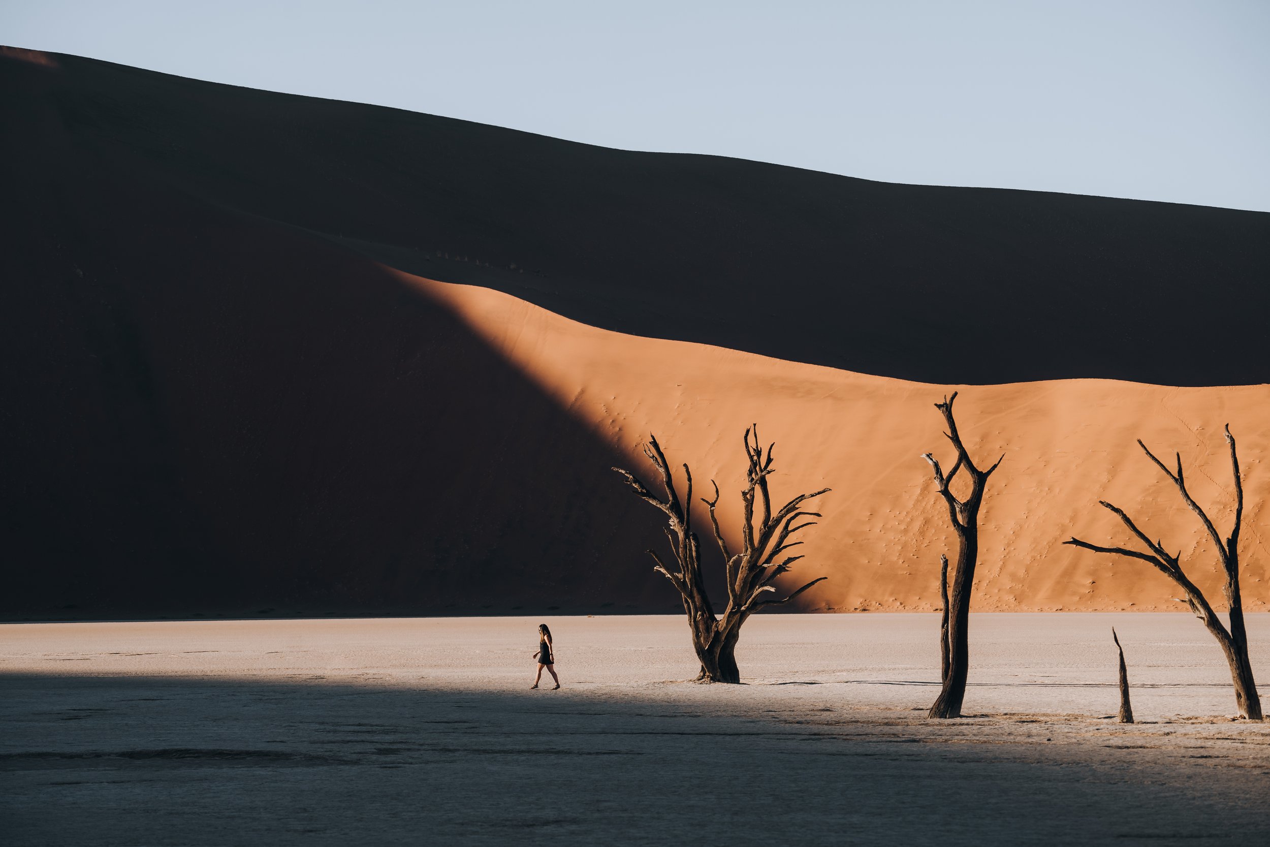A lone person walking across the white clay pan at Deadvlei in Namibia, flanked by skeletal dead acacia trees, with a dramatic sand dune and dark mountain in the background under clear light.
