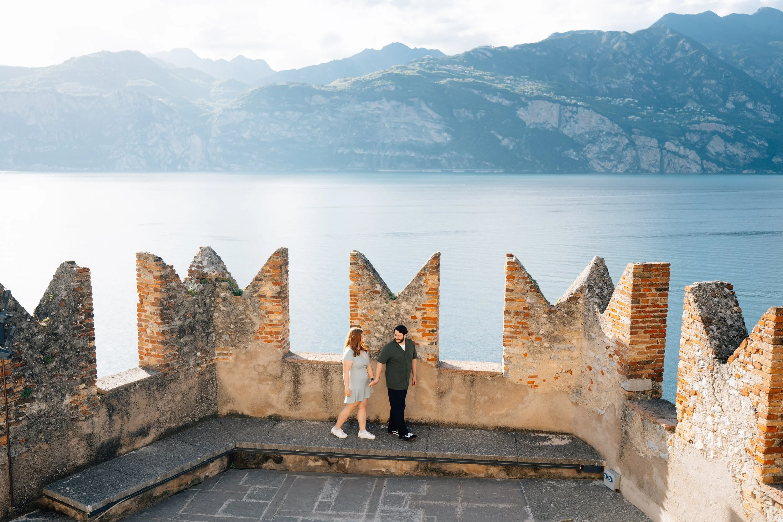 A couple standing on the stone terrace of Castello Scaligero in Malcesine overlooking the blue waters and mountains of Lake Garda