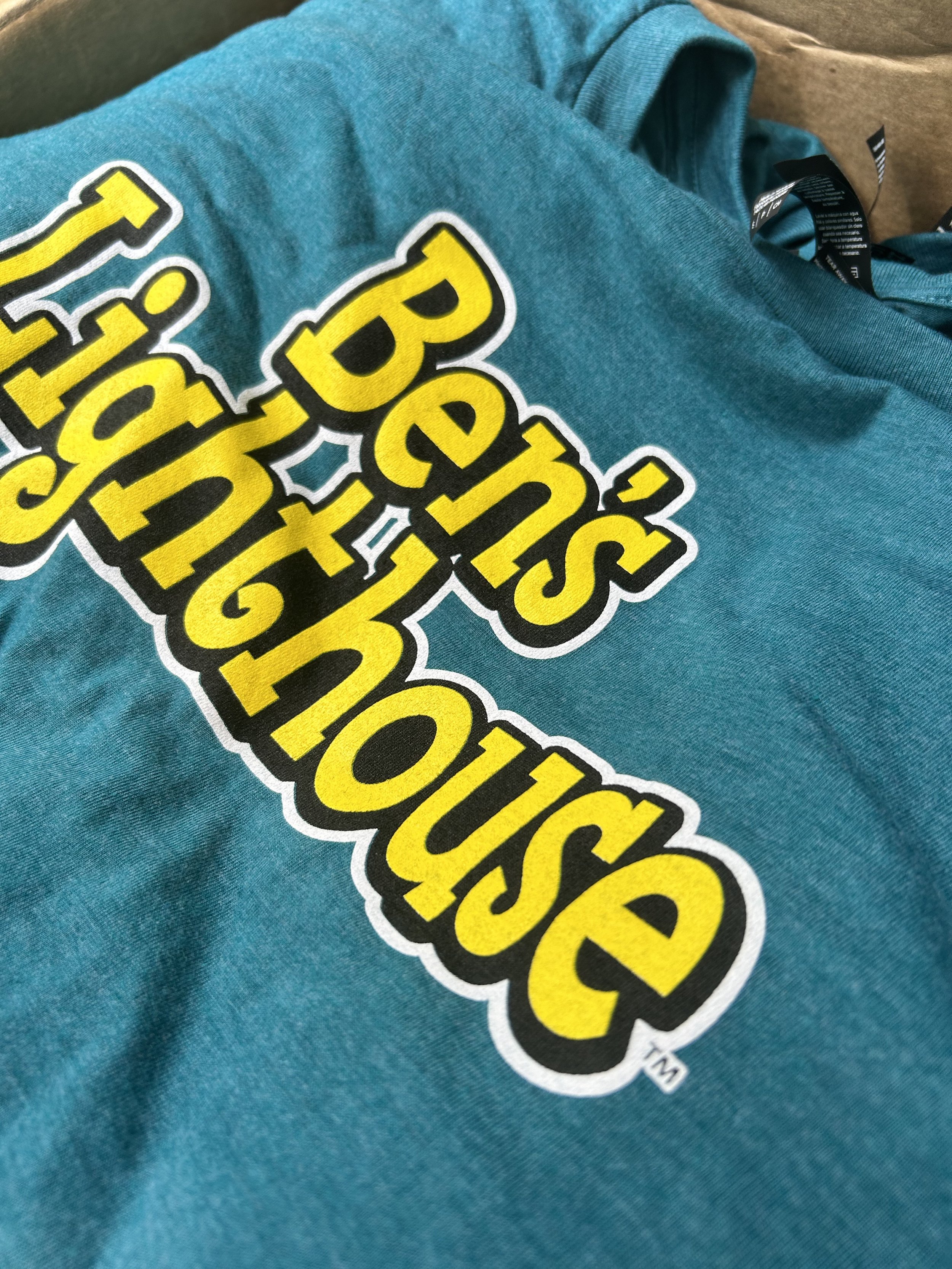 Close-up of a blue T-shirt with the text 'Ben's Lighthouse' in bold yellow and black screen printed ink.