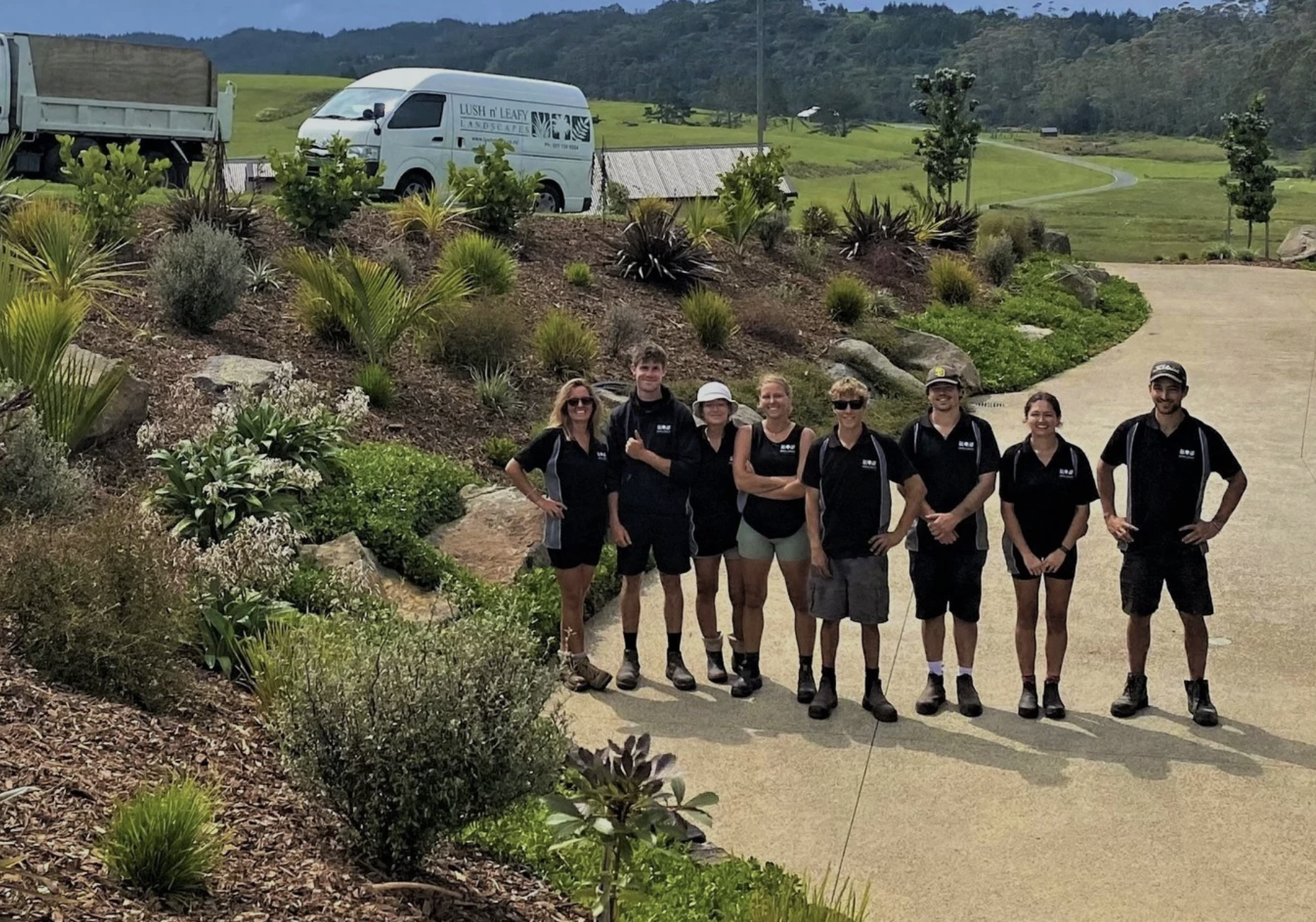 A group of eight people standing on a cement pathway in a landscaped garden area with various plants and rocks, smiling at the camera, with a hilly landscape in the background, a white van reading 'Lush n Leafy Landscapes,' and a large truck parked nearby.