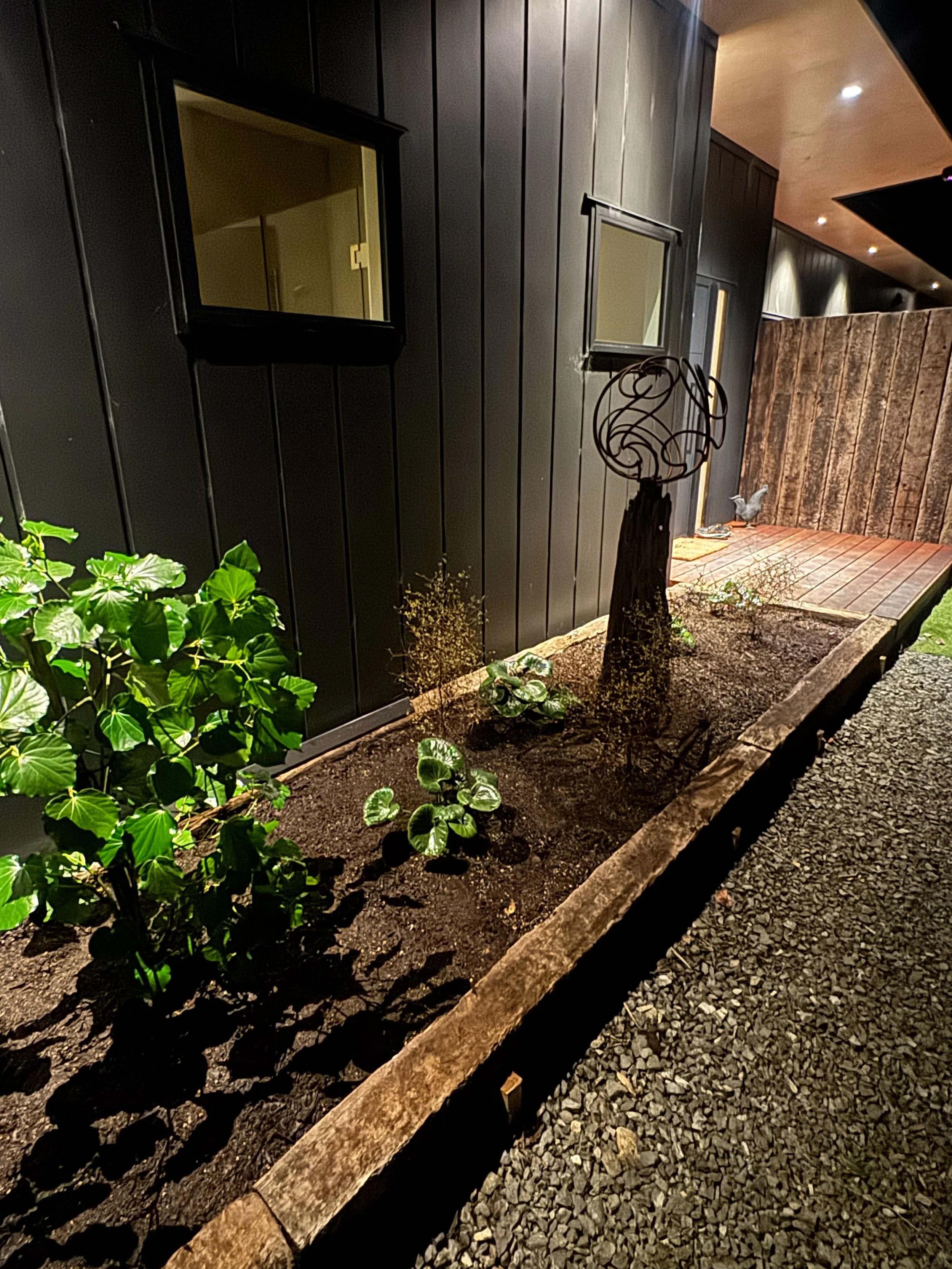 Nighttime outdoor scene showing a small garden bed with green plants, a black decorative garden ornament, and a potted plant. The garden bed is bordered by a weathered wooden plank, with gravel surrounding it. In the background, a dark wooden wall an