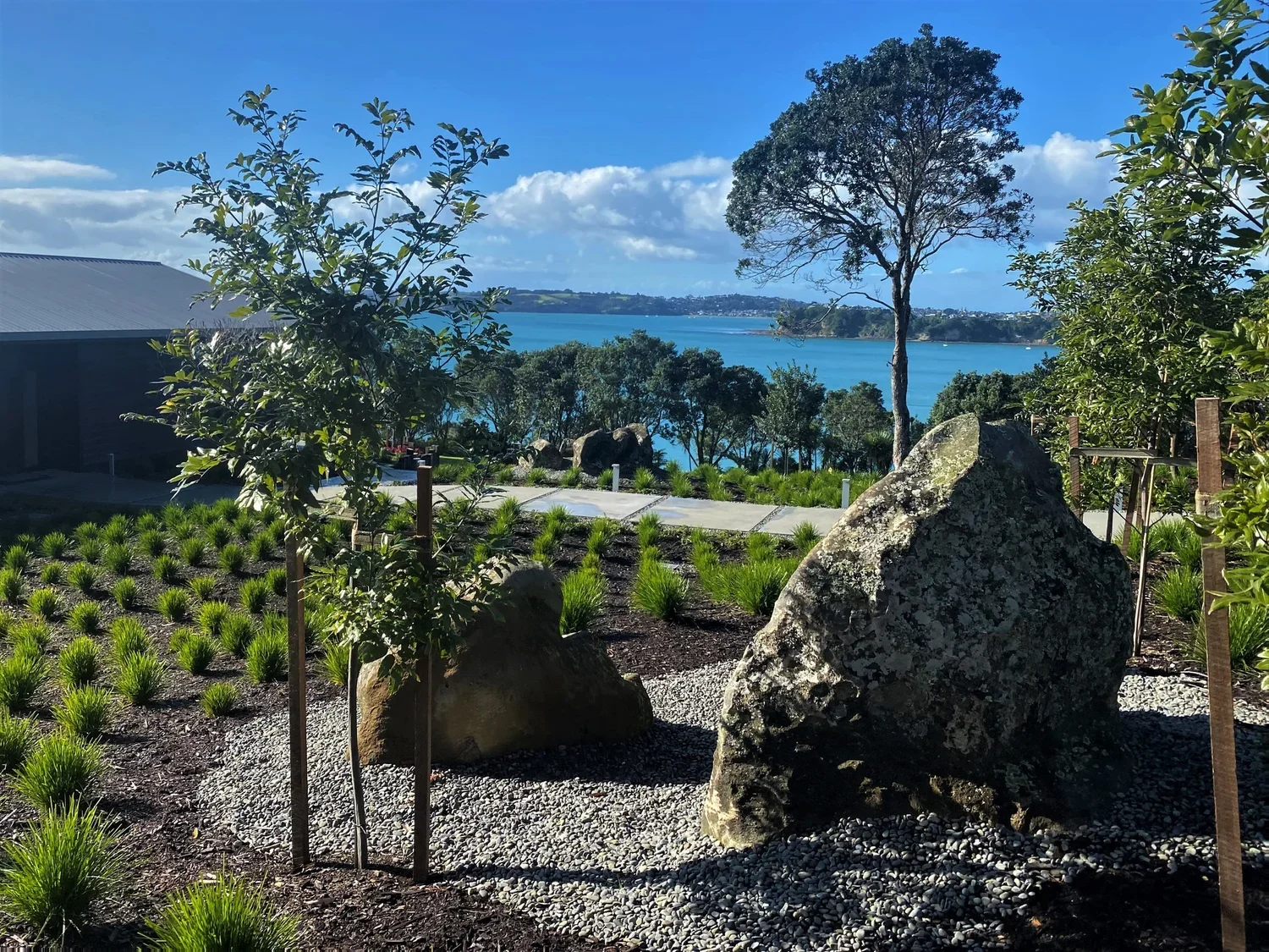 Landscape view of a garden with large rocks, green shrubs, and a tree, overlooking a water body and distant shoreline under a partly cloudy sky.