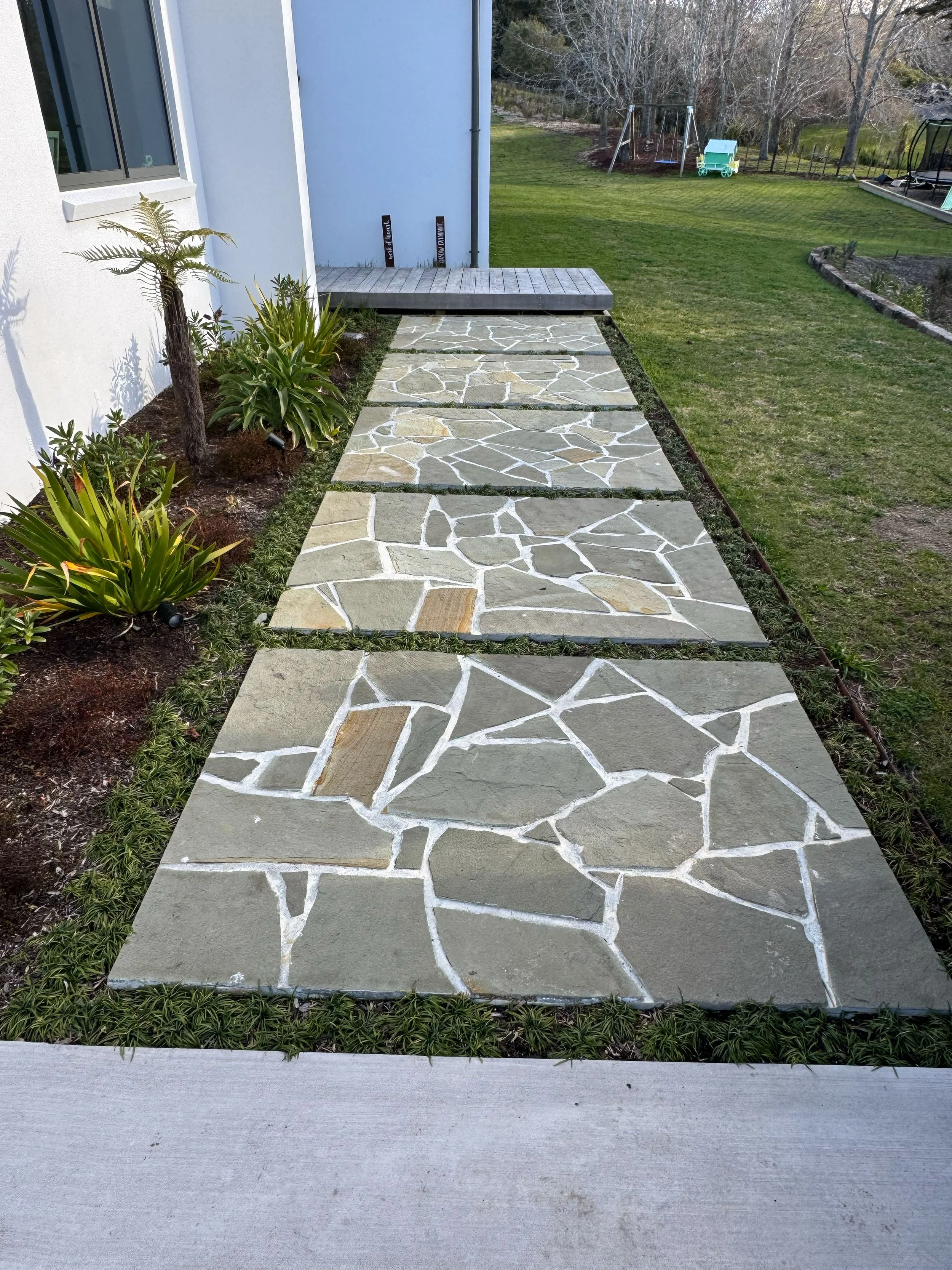 View of a stone pathway with large rectangular and irregular pieces, bordered by a flower bed, leading up to a back door on a white house. A small deck is at the end of the pathway, with two signs leaning against the house wall. In the background, th