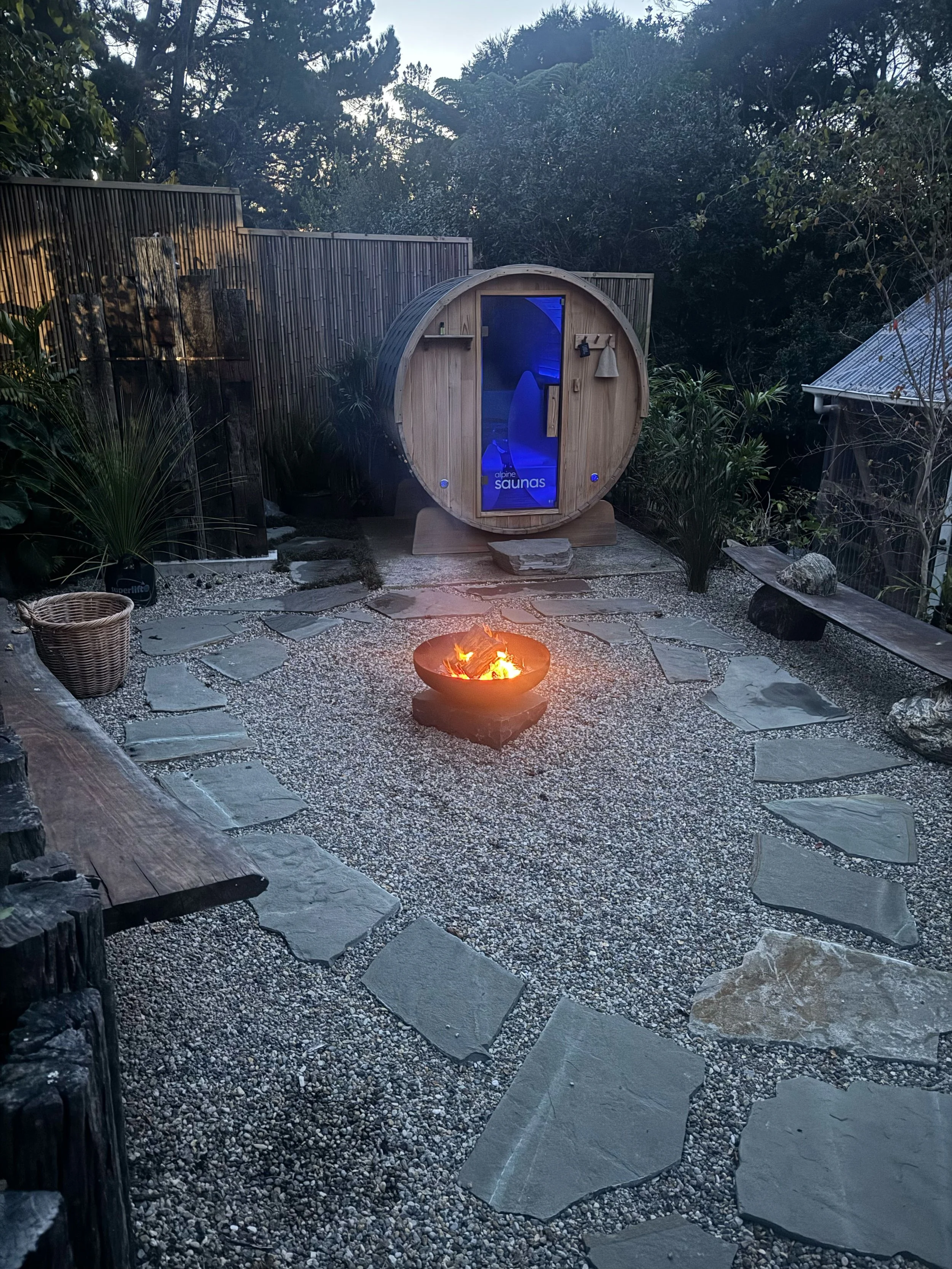 Outdoor backyard with a fire pit, stone pathway, wooden Bench, sauna cabin, and lush trees and plants.