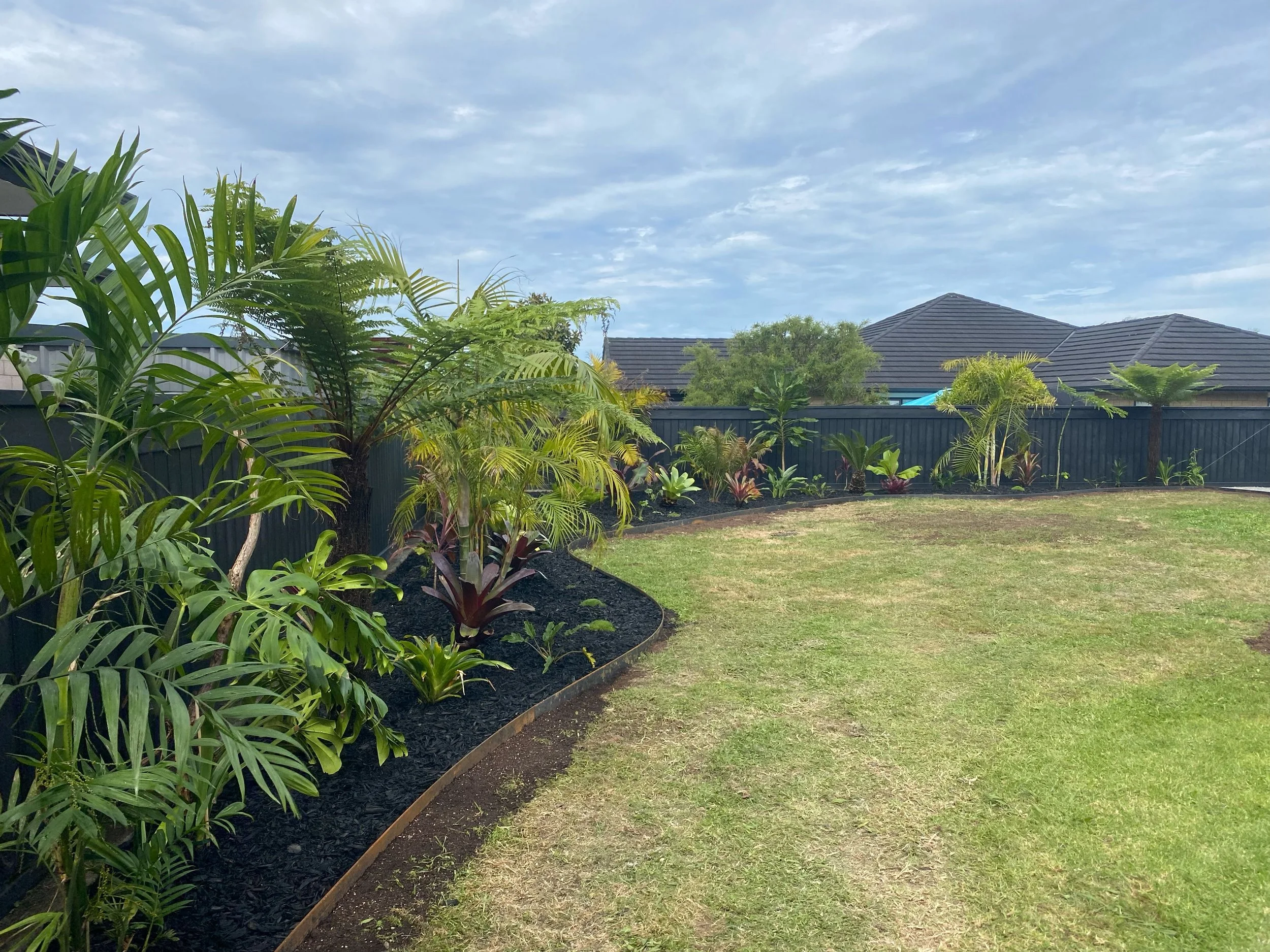 Backyard garden with various green plants and trees, a black fence, and neighboring houses with dark roofs under a cloudy sky.