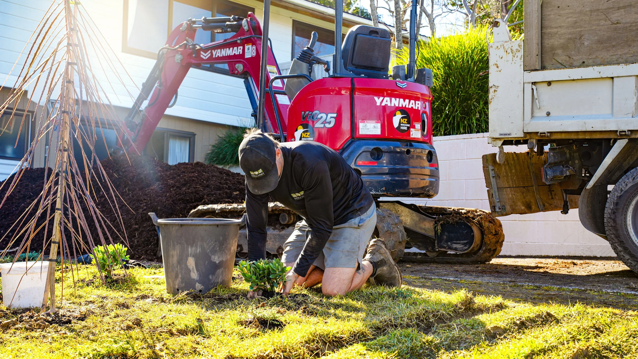 A person is planting a small shrub in a garden with a red Yanmar mini excavator behind them, near a light-colored house, on a sunny day.
