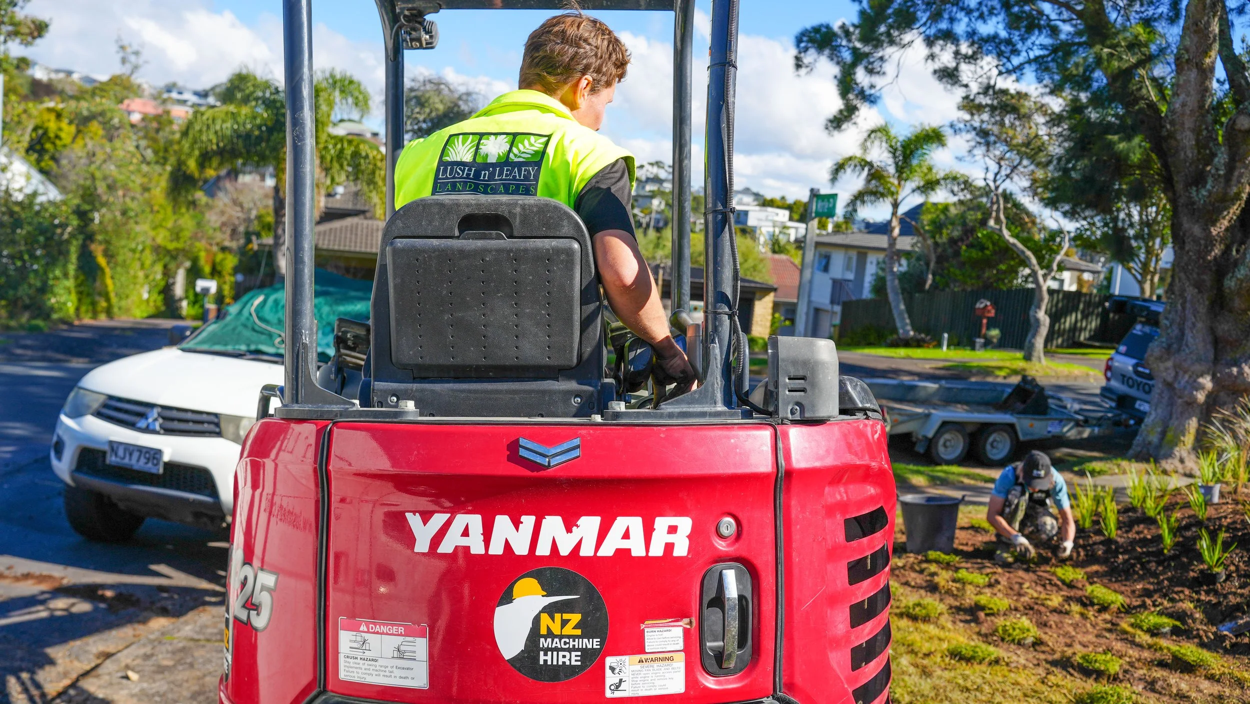A person wearing a yellow vest with a landscaped logo operates a red Yanmar mini excavator on a residential street. Behind, another person is working in a garden bed, with trees and houses in the background under a blue sky with scattered clouds.