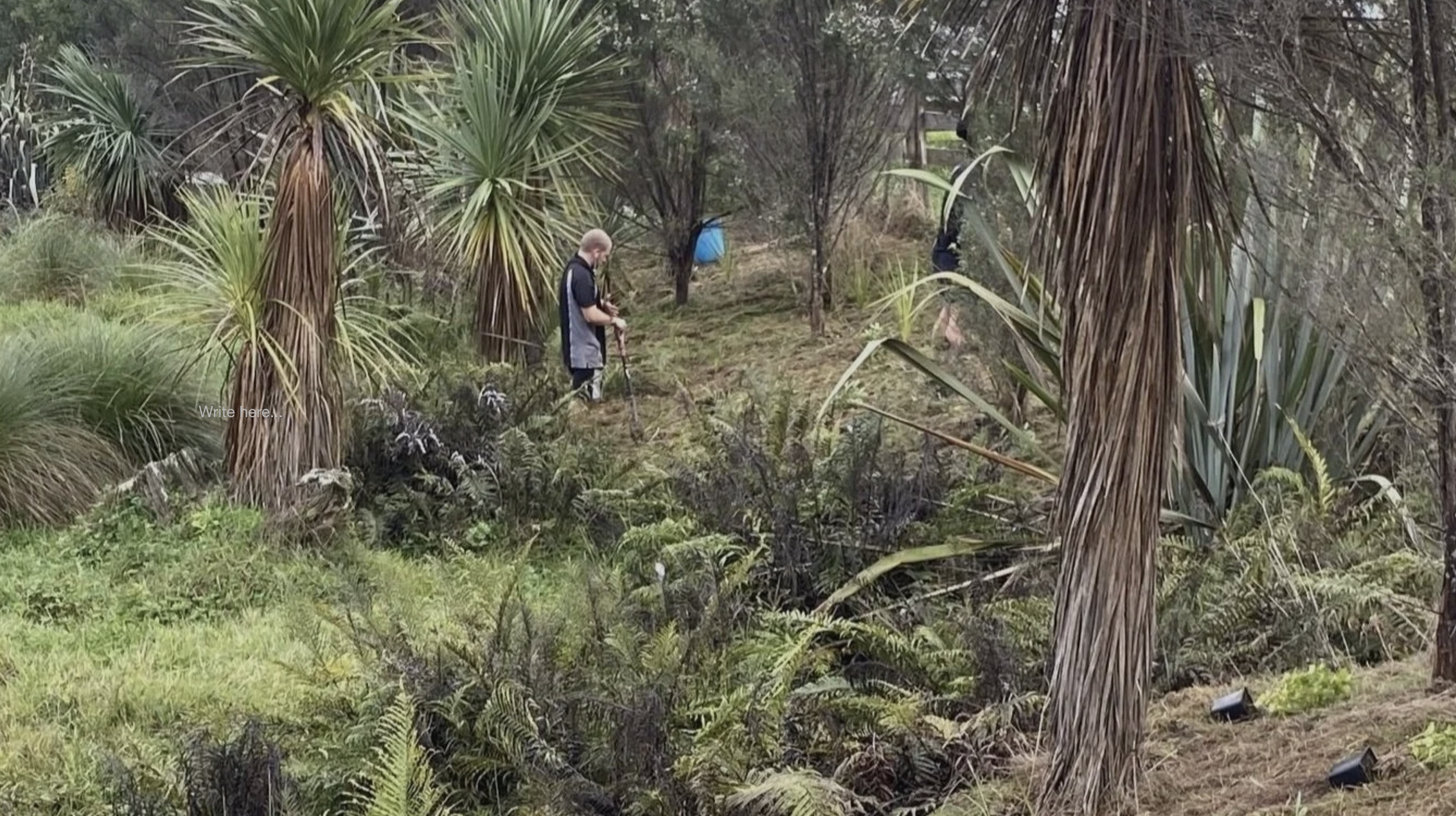 A person with light hair dressed in dark clothing standing in a lush, green garden surrounded by tall, spiky plants and trees, with another person partially obscured in the background.