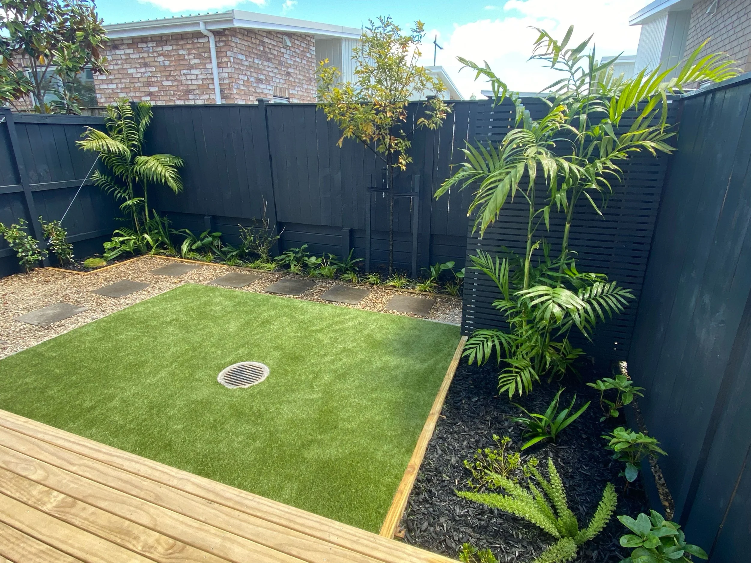A backyard with a small artificial grass lawn, a wooden deck, a gravel pathway, and a black privacy fence with various green plants and trees.