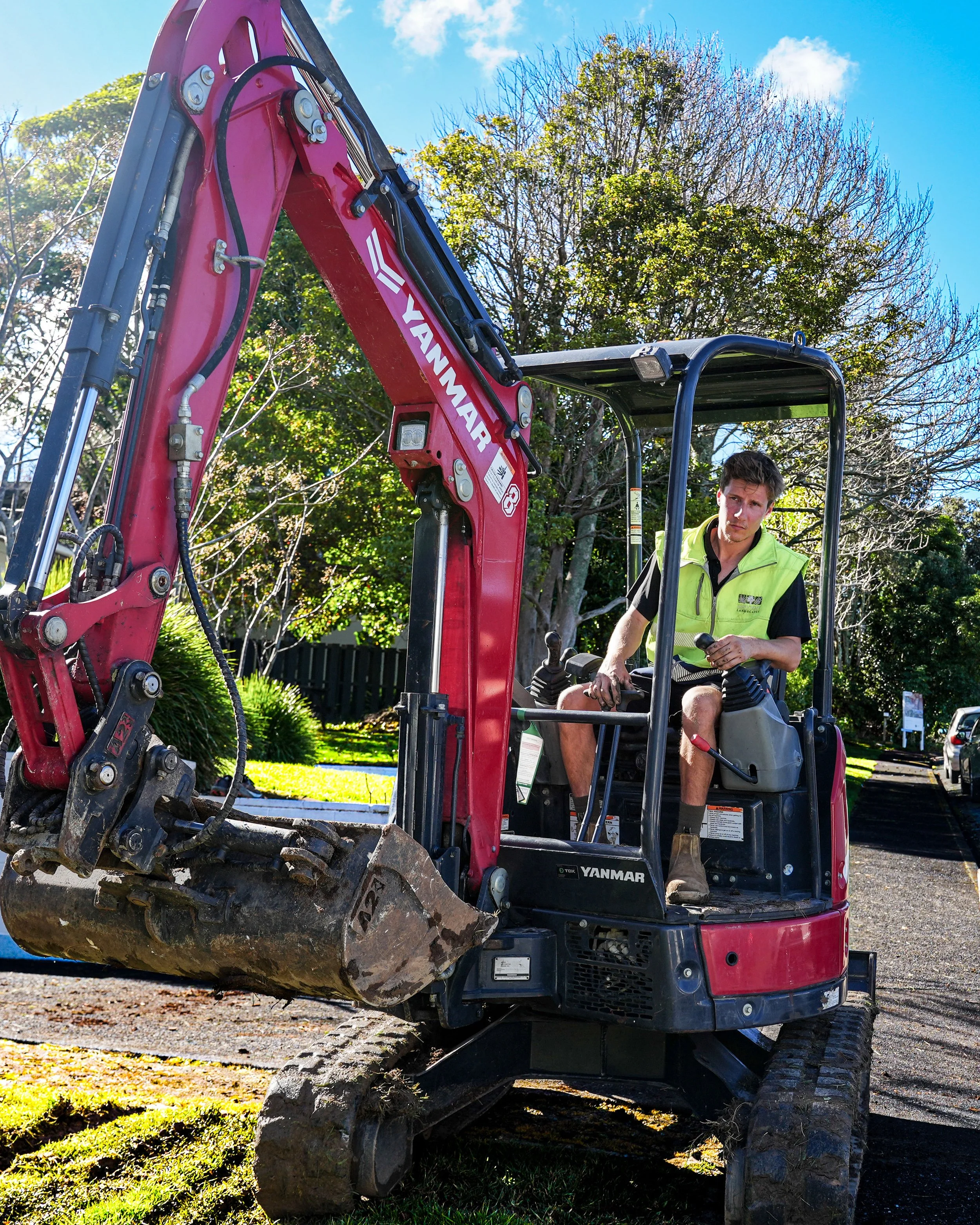 A man operating a red Yanmar compact excavator with large dirt bucket, outdoor setting with trees and blue sky.