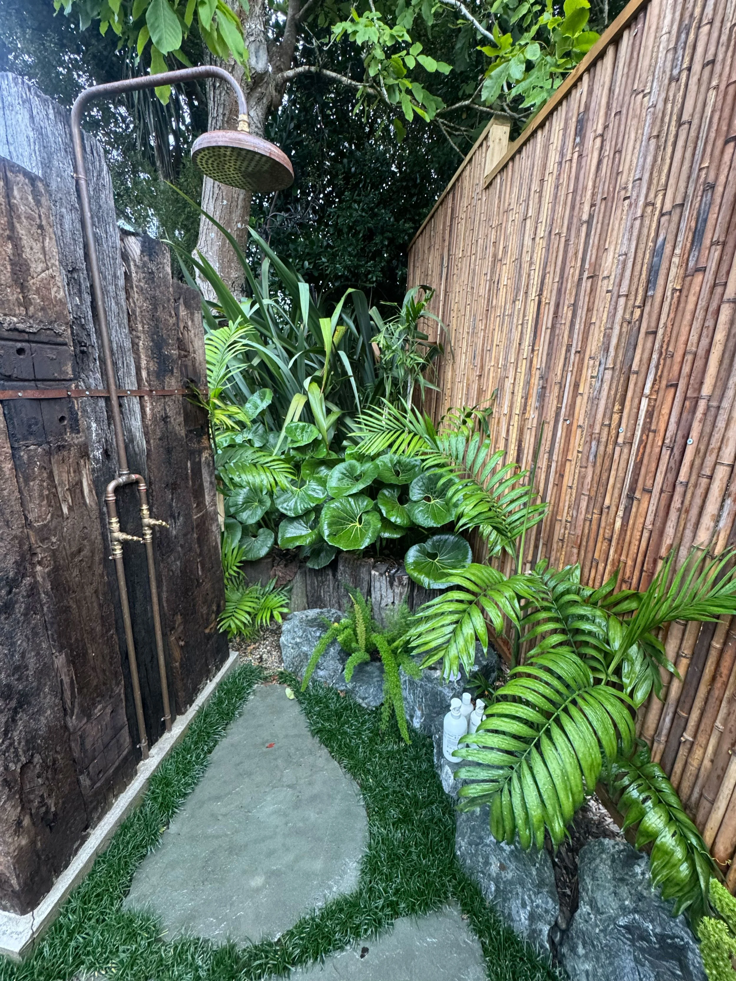 A small outdoor shower area with a wooden and bamboo fence, lush green plants, and large rocks, with bottles of soap or lotion placed on the ground.
