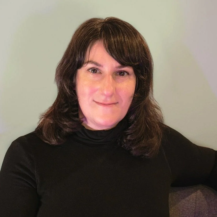 A woman with brown hair wearing a black turtleneck, smiling at the camera in front of a pale green wall.