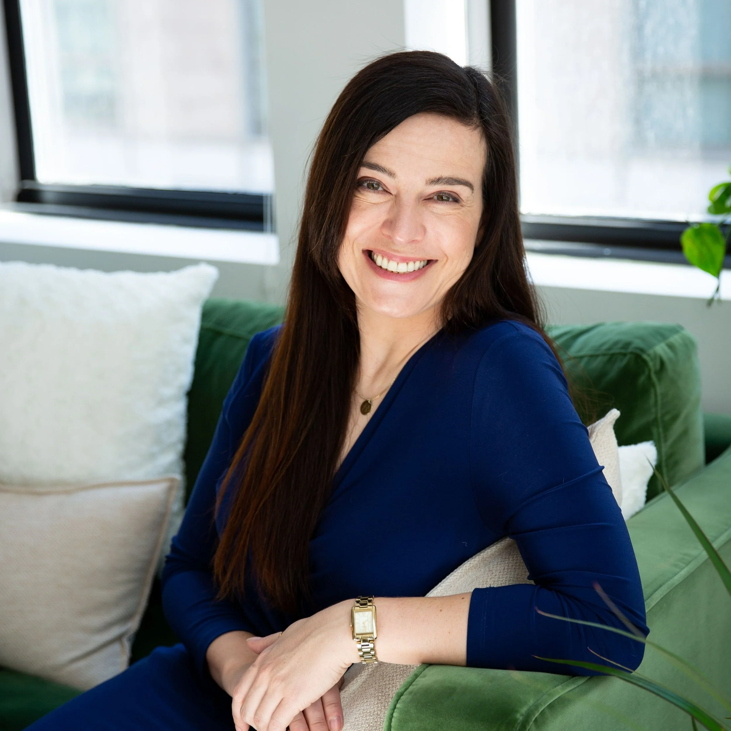 A woman with long dark hair, wearing a blue top, sitting on a green sofa with white pillows, smiling and looking at the camera, in a bright room with large windows.