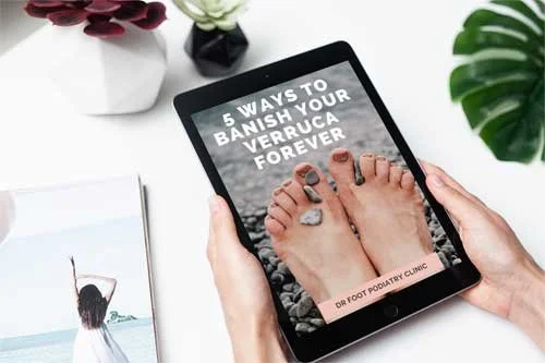 A person's hands holding a tablet displaying a guide titled '5 Ways to Banish Your Stress Forever' with an image of feet with stones on toes, on a desk with a plant, flowers, and a magazine.