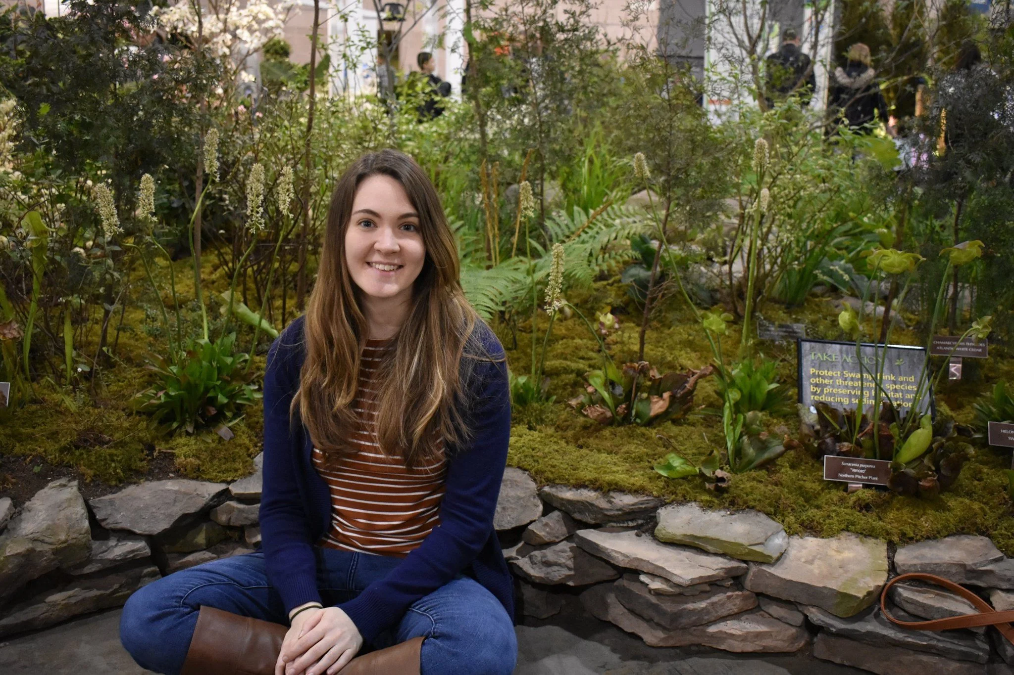 A white woman with long brown hair sits cross legged in front of a stone wall and lots of plants.