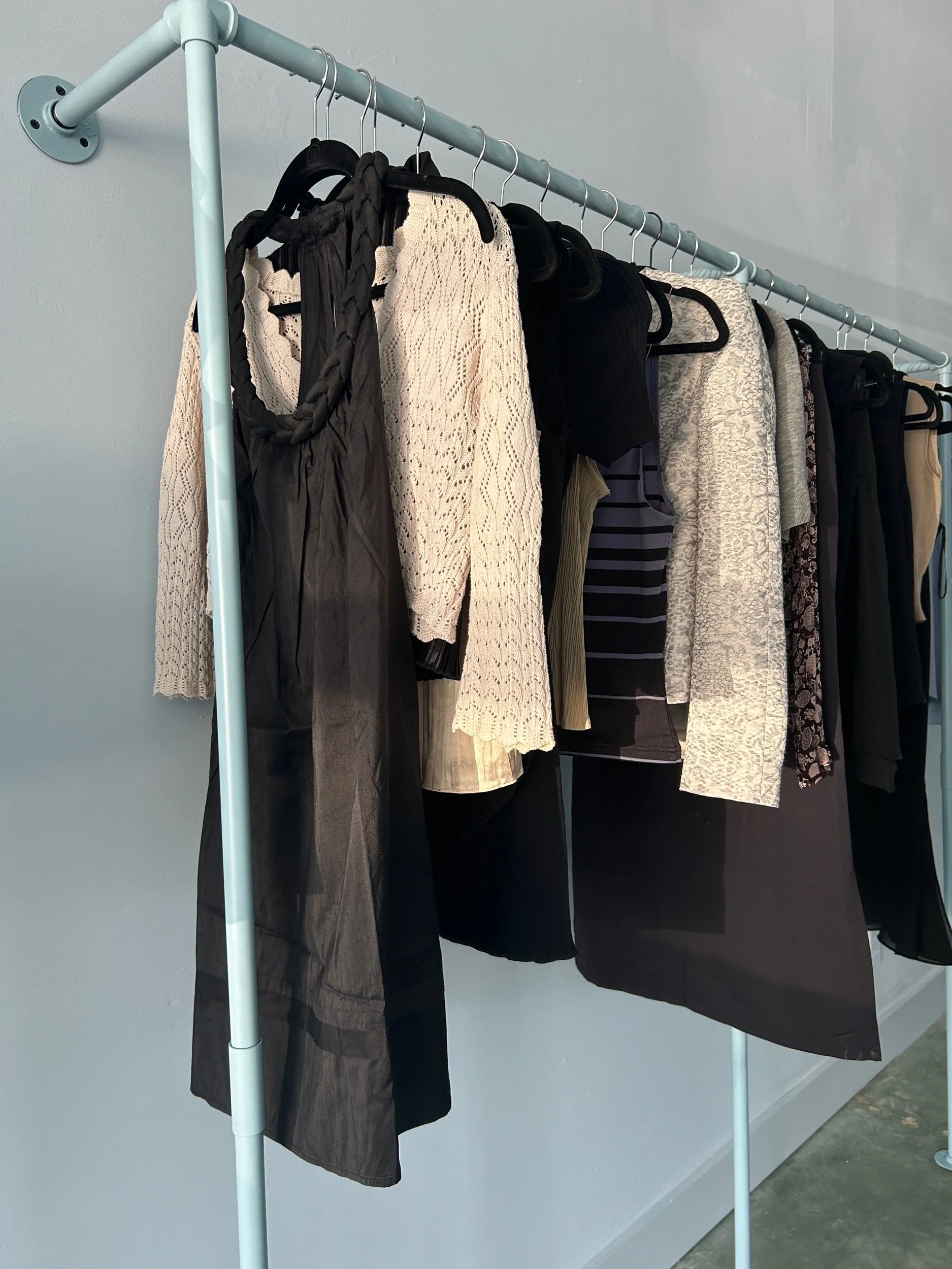 Clothing rack with dresses, skirts, and tops hanging on black hangers against a gray wall.
