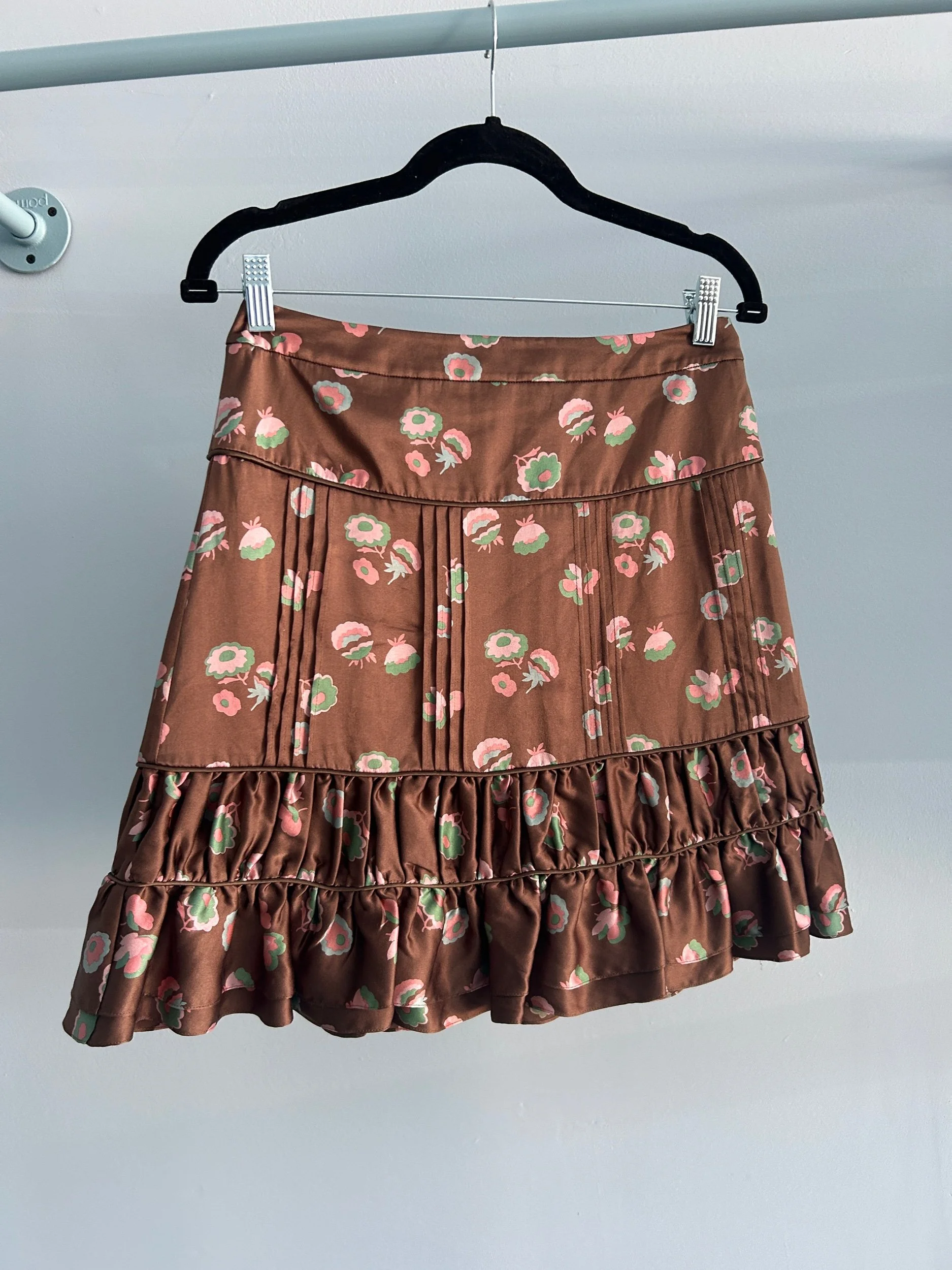 A March Jacobs silk brown skirt with a pink and green floral pattern hanging on a black hanger, attached to a clothing line with clips.