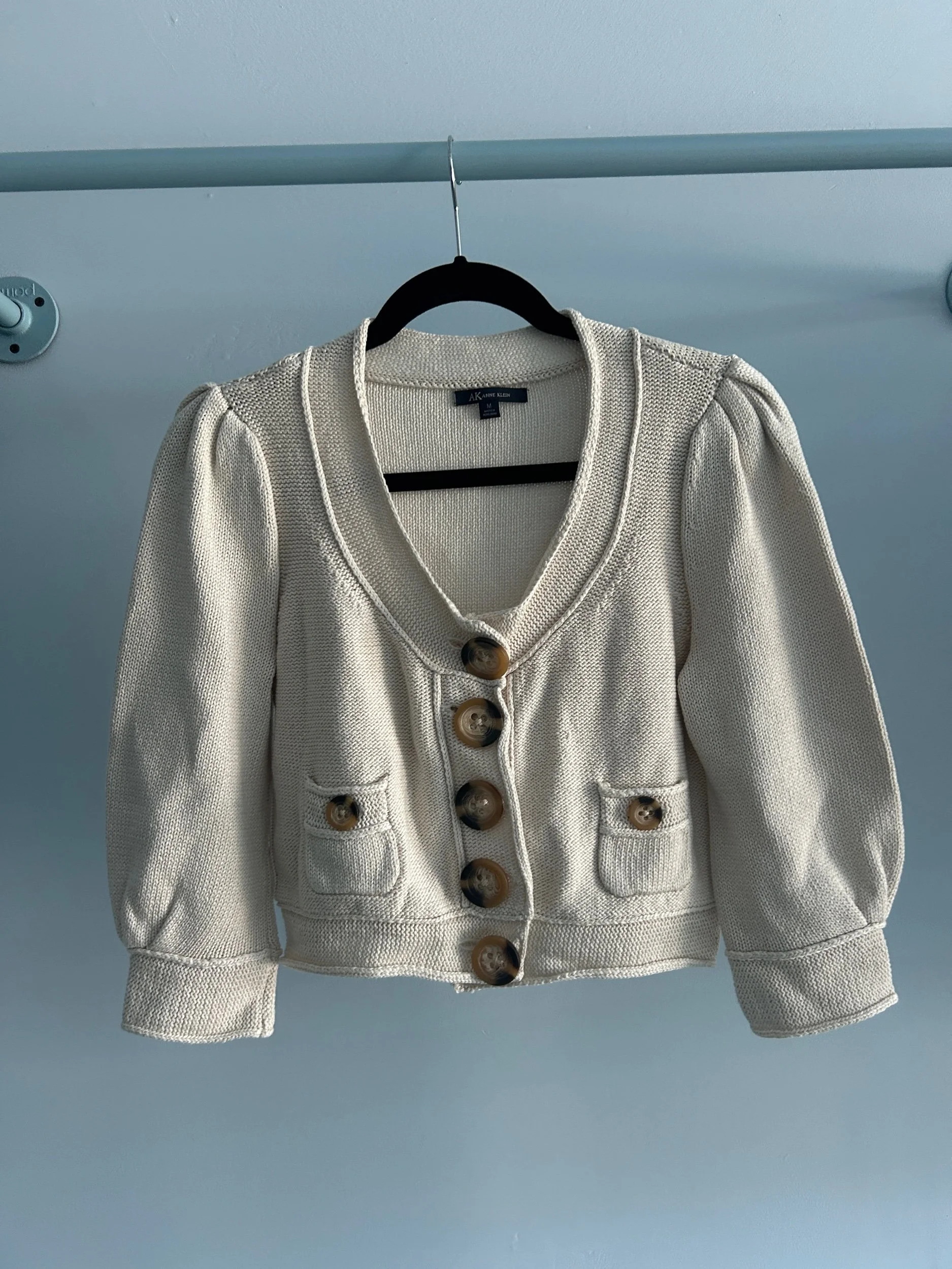 A cream-colored knitted sweater with large brown buttons hanging on a black hanger against a light blue wall.