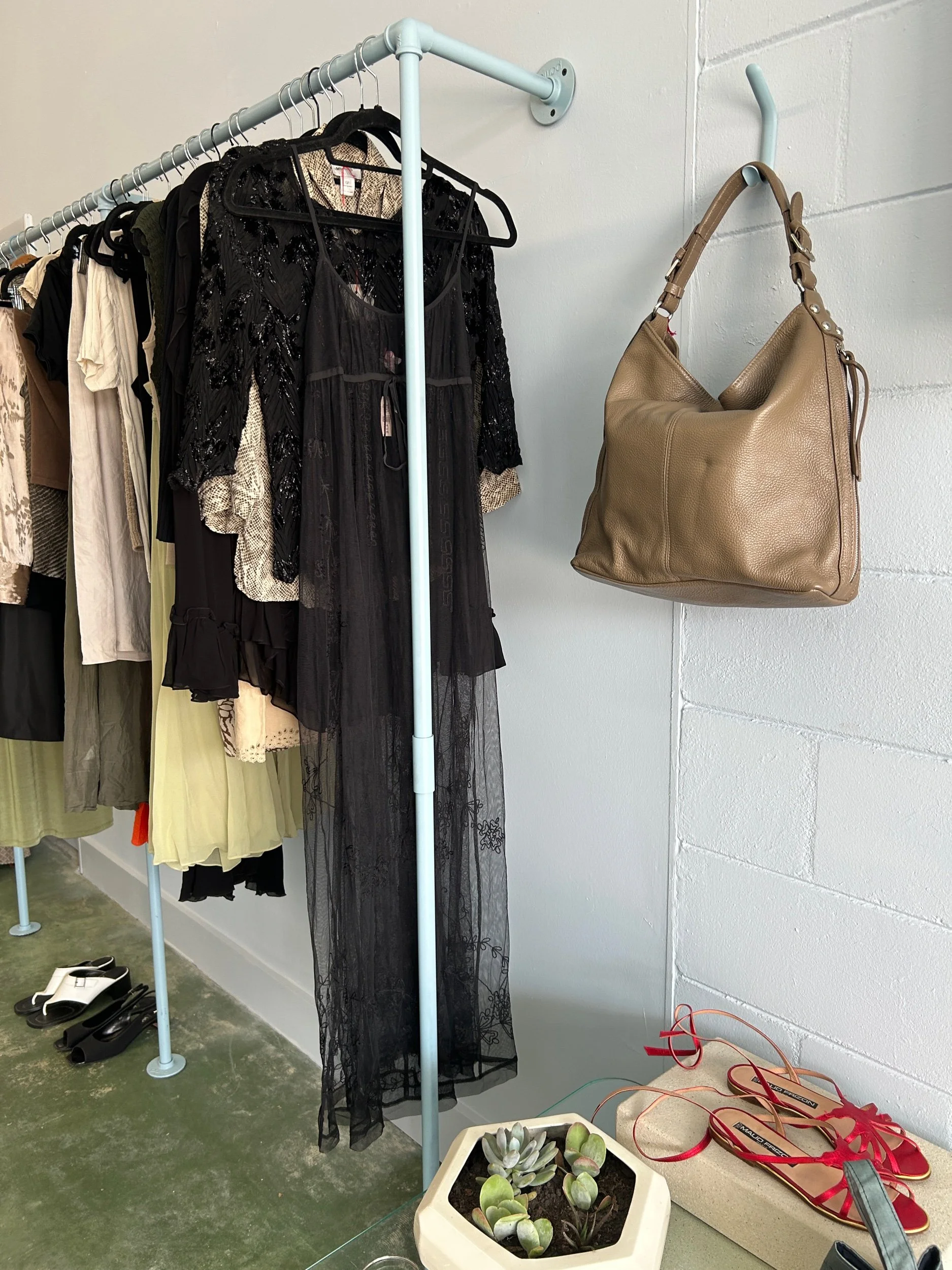 Clothing rack with black and beige dresses, black and white shoes on the floor, a beige handbag hanging on the wall, and a plant pot with succulents on a glass table.