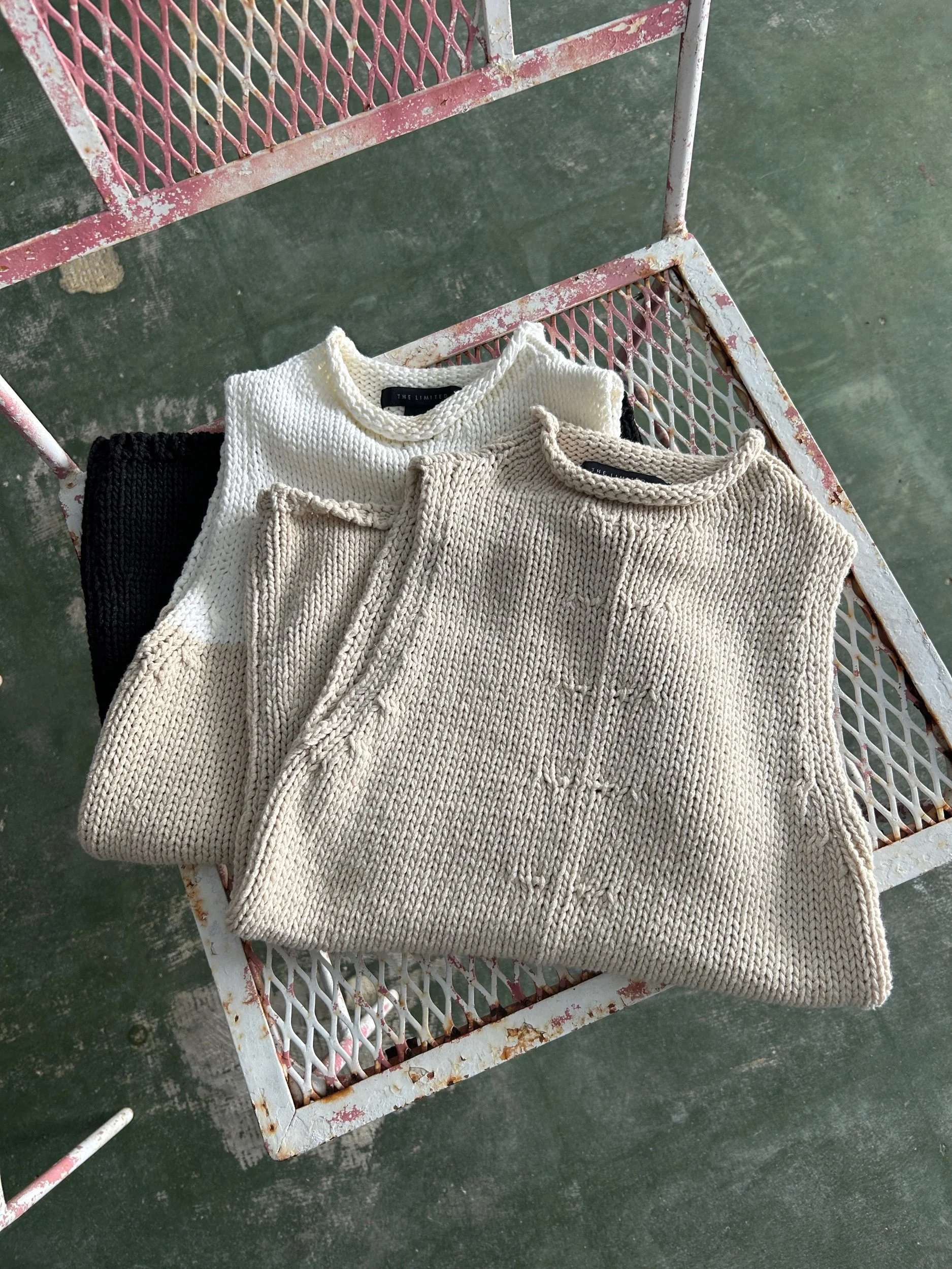 A rusty white metal cart with pink accents, holding three folded knitted sweaters in black, white, and beige, placed on a green concrete floor.