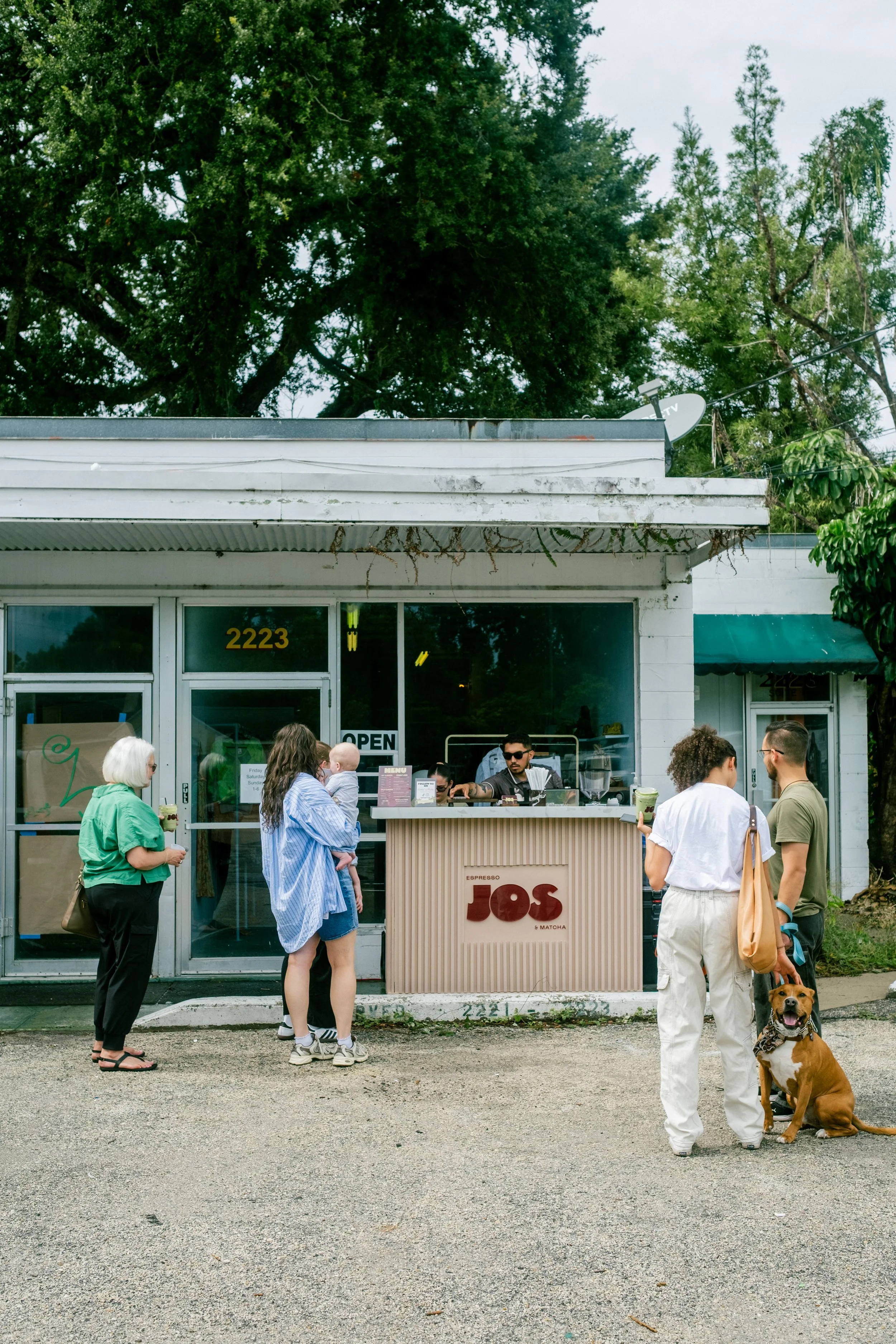 People gather outside a small cafe or shop with a sign that reads "JOS." Several individuals are standing and chatting, with one person sitting behind the counter. A large dog sits on the ground in front of a person with curly hair. Trees are visible in the background, and the building has a white facade with large windows.