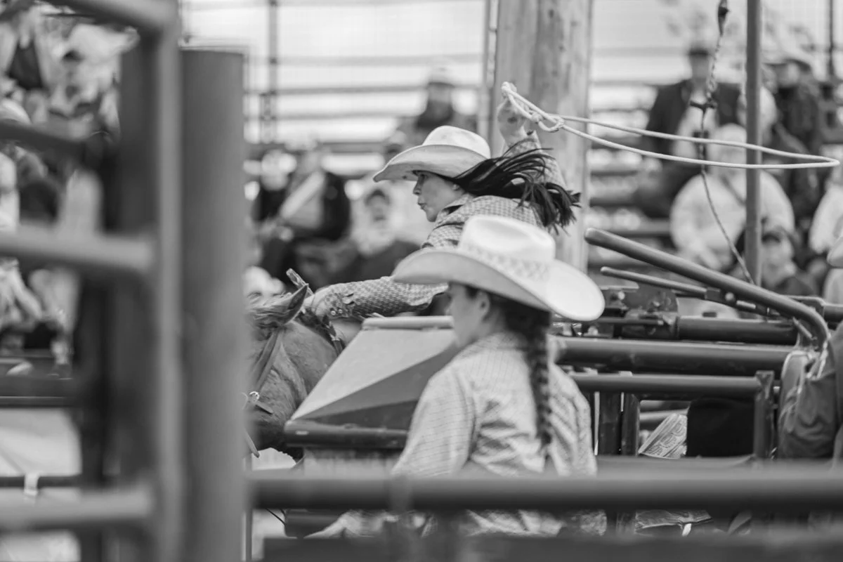 A young woman is on her horse running forward into the arena as she tosses her rope up in the air behind her for breakaway roping at the Brash Rodeo. Around her, you can see all of the spectators and other cowgirls awaiting their turn. 