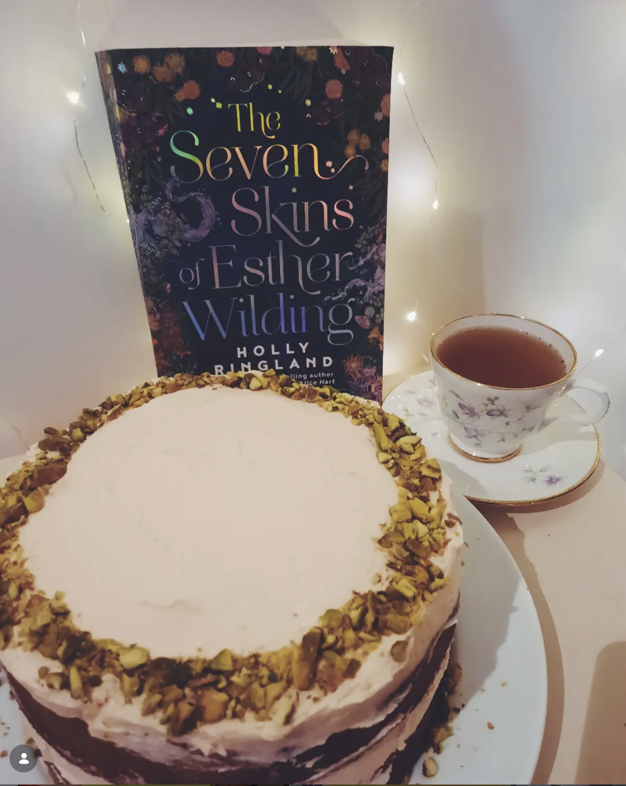 A cake with white icing and chopped nuts around the top edge, a cup of tea in a floral teacup and saucer, and a hardcover book titled 'The Seven Skins of Esther Wilding' by Holly Ringland in the background with fairy lights.