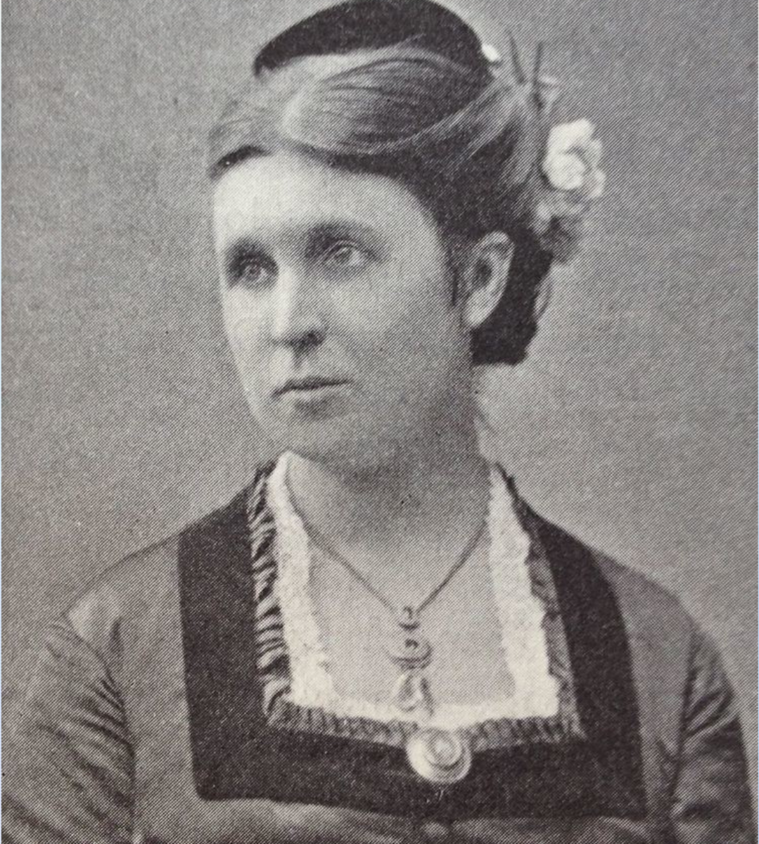 Black-and-white portrait of a woman with a side bun hairstyle, wearing a dress with lace detailing and decorative pins, and a necklace with a pendant.