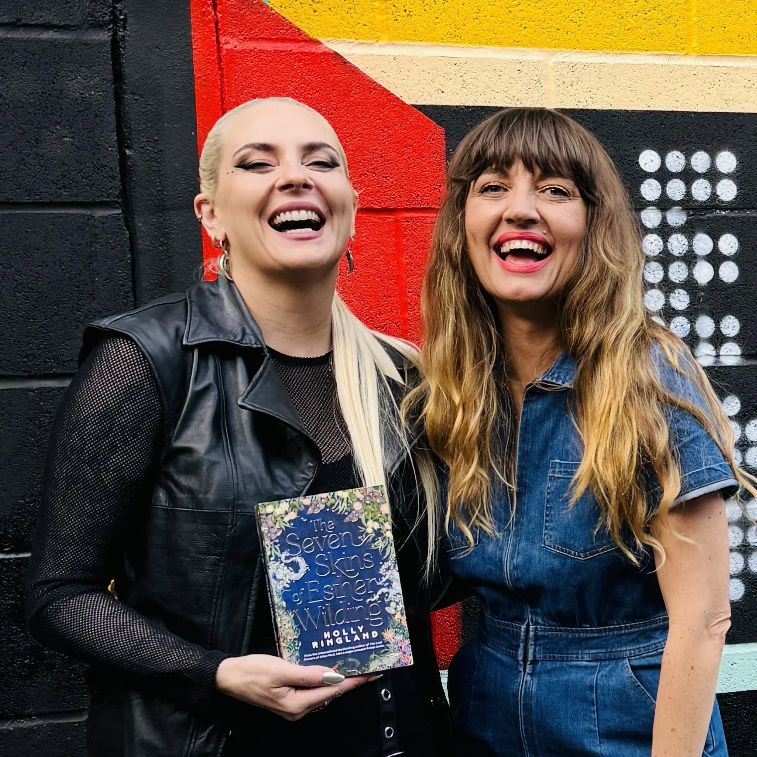 Two women smiling and laughing in front of a colorful mural. One woman holds a book titled 'The Seven Skins of Esther Wullow' by Holly Ringland.
