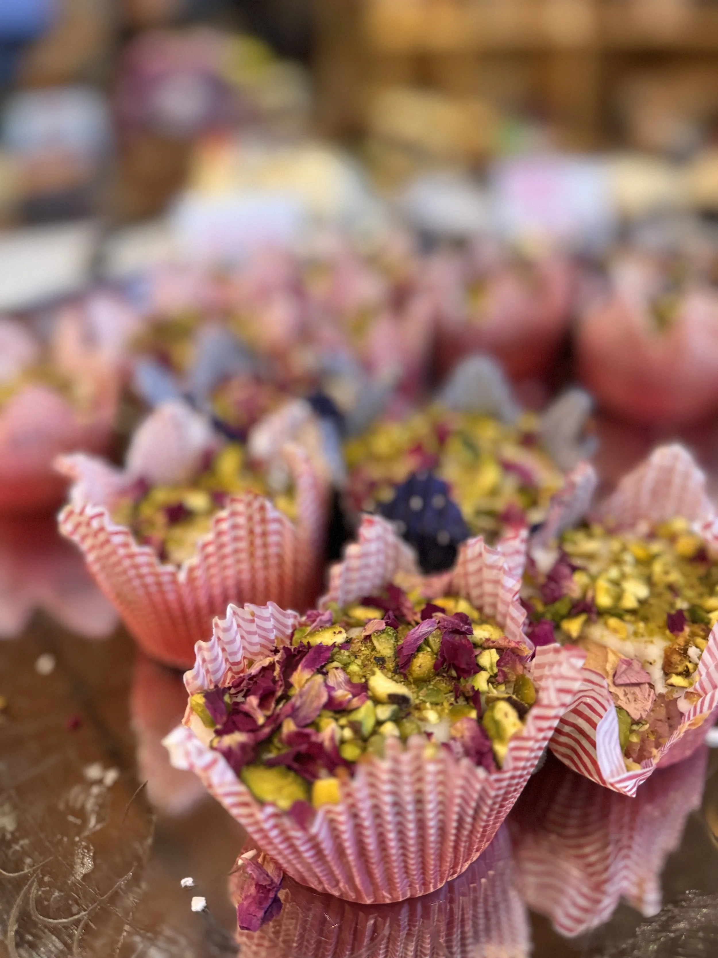 Colorful candies topped with chopped pistachios, wrapped in pink and white striped paper, arranged in a display.