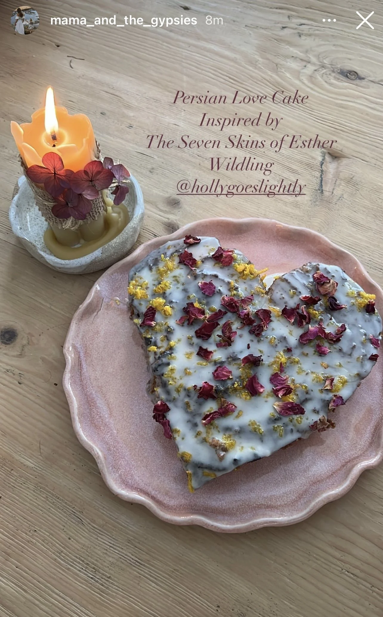 Heart-shaped Persian love cake with white glaze and sprinkled with dried rose petals and yellow flower petals on a pink ceramic plate, next to a lit candle decorated with dried flower petals and leaves on a wooden surface.