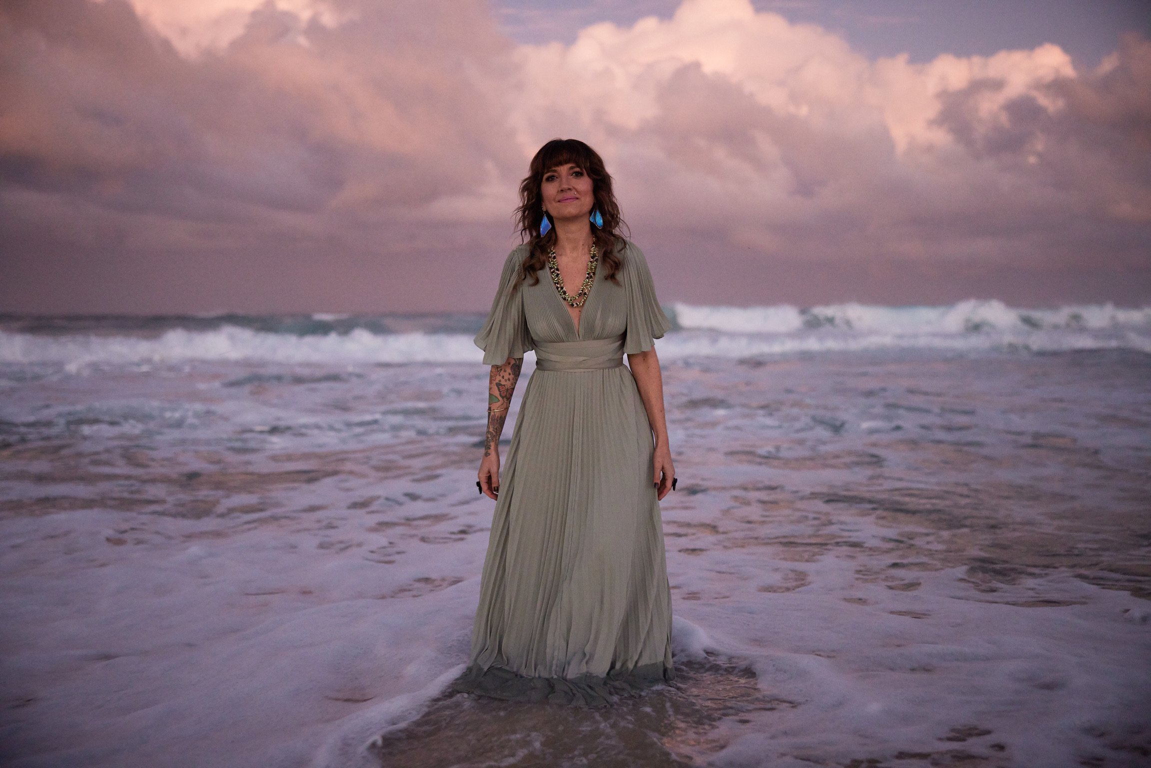 A woman in a long beige dress with blue earrings stands in the ocean with waves at her feet, under a pinkish-purple cloudy sky.