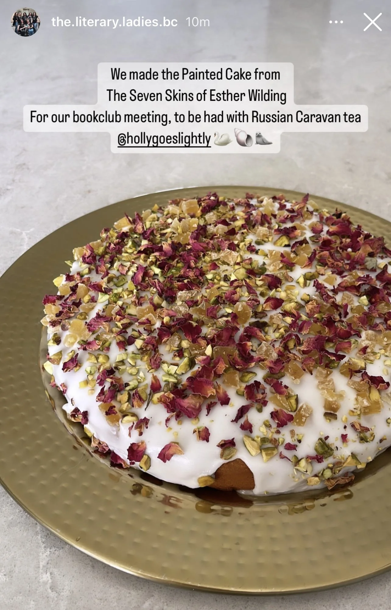 A painted cake covered with white frosting, topped with dried rose petals, chopped pistachios, and small pieces of candied fruit, sitting on a gold plate.