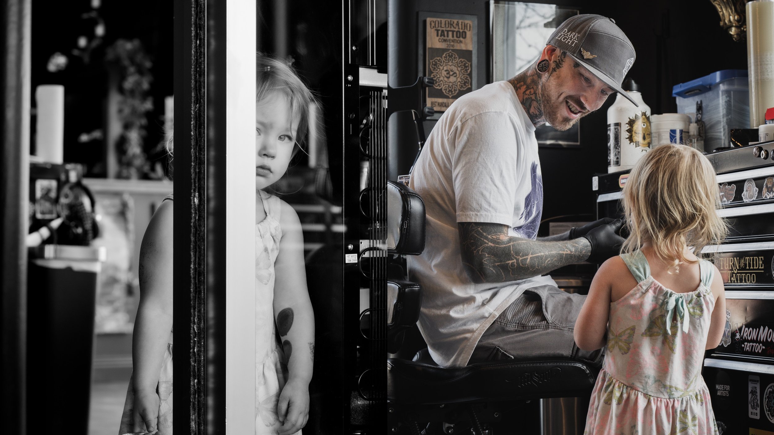 Zach Atlan's daughter, Willow, peers through the shop window during a candid portrait on Apr. 16, 2022, in Fort Collins, Colo. For Atlan, owning Dark Light is an extension of his immediate family. "This shop is my tribe," Atlan said. "I love the art.